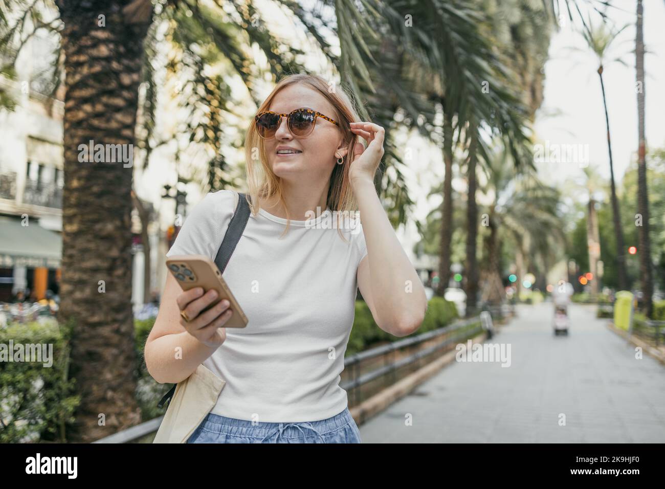 Primo piano di una giovane donna felice che guarda il suo smartphone mentre cammina per strada. Ragazza Millennial che utilizza app mobile all'aperto. Foto Stock
