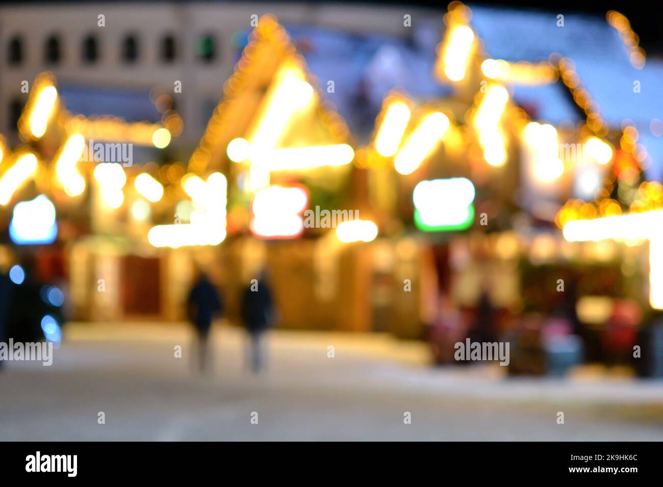 Sfondo sfocato. La gente cammina nella piazza della città la sera d'inverno. Silhouette nere di persone che camminano vicino a case decorate illuminazione luminosa. Luce bianca bokeh sfocare i punti dalle luci della casa incandescente Foto Stock