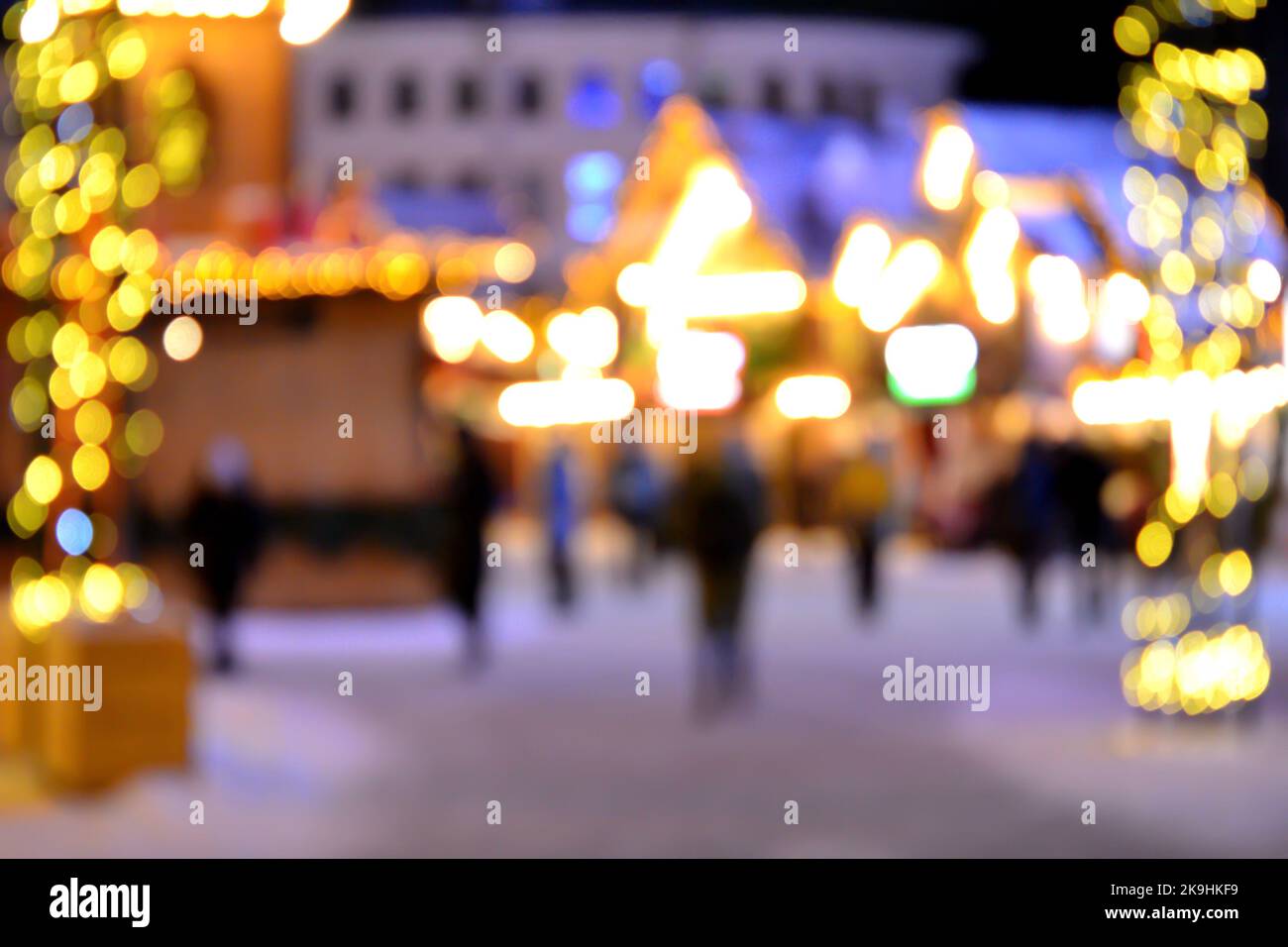 Sfondo sfocato. La gente cammina nella piazza della città la sera d'inverno. Silhouette nere di persone che camminano vicino a case decorate illuminazione luminosa. Luce bianca bokeh sfocare i punti dalle luci della casa incandescente Foto Stock