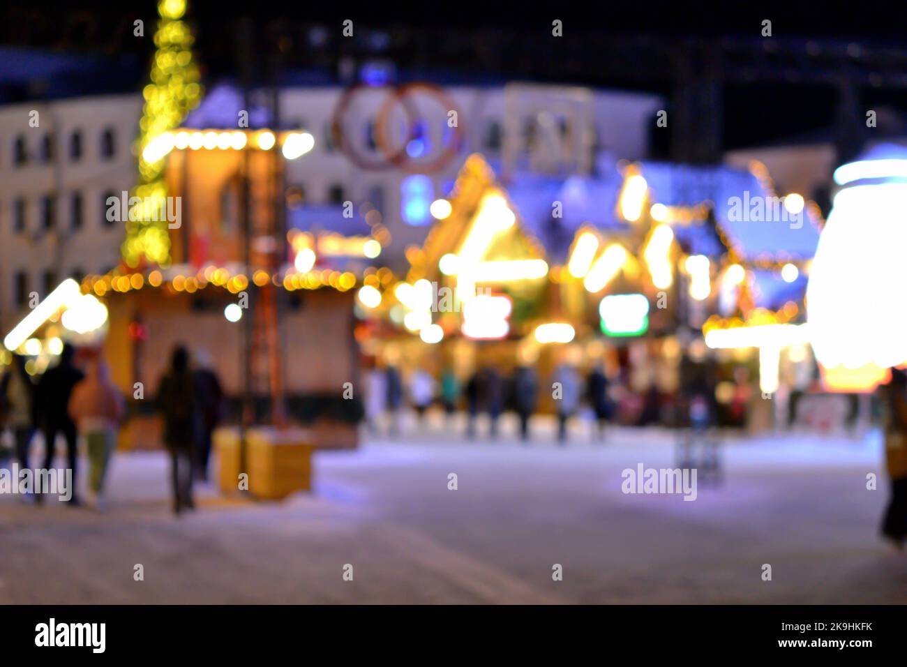 Sfondo sfocato. La gente cammina nella piazza della città la sera d'inverno. Silhouette nere di persone che camminano vicino a case decorate illuminazione luminosa. Luce bianca bokeh sfocare i punti dalle luci della casa incandescente Foto Stock
