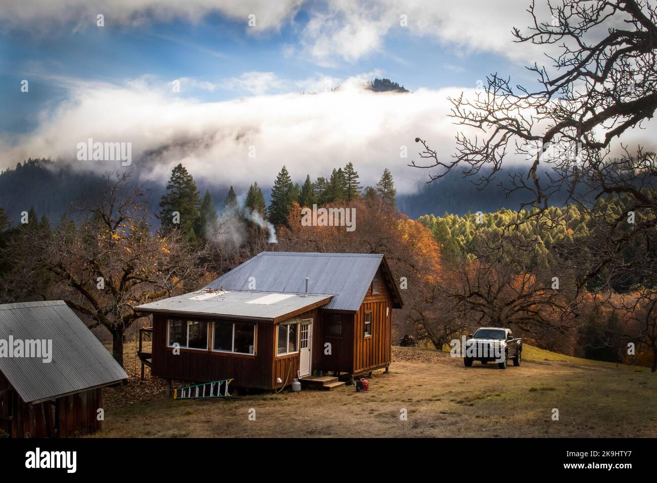 Cabina in legno nelle montagne nebbiose all'ora d'oro con fumo che sale da stovepipe e camion parcheggiato all'esterno Foto Stock