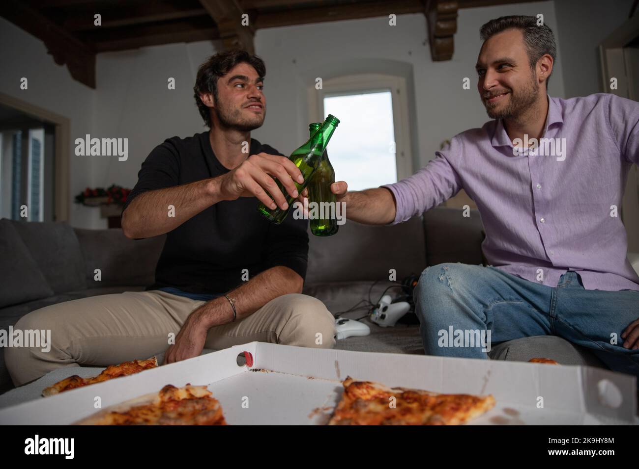 Due amici brindano con due birre in pausa dal videogioco che mangia la pizza. Foto Stock