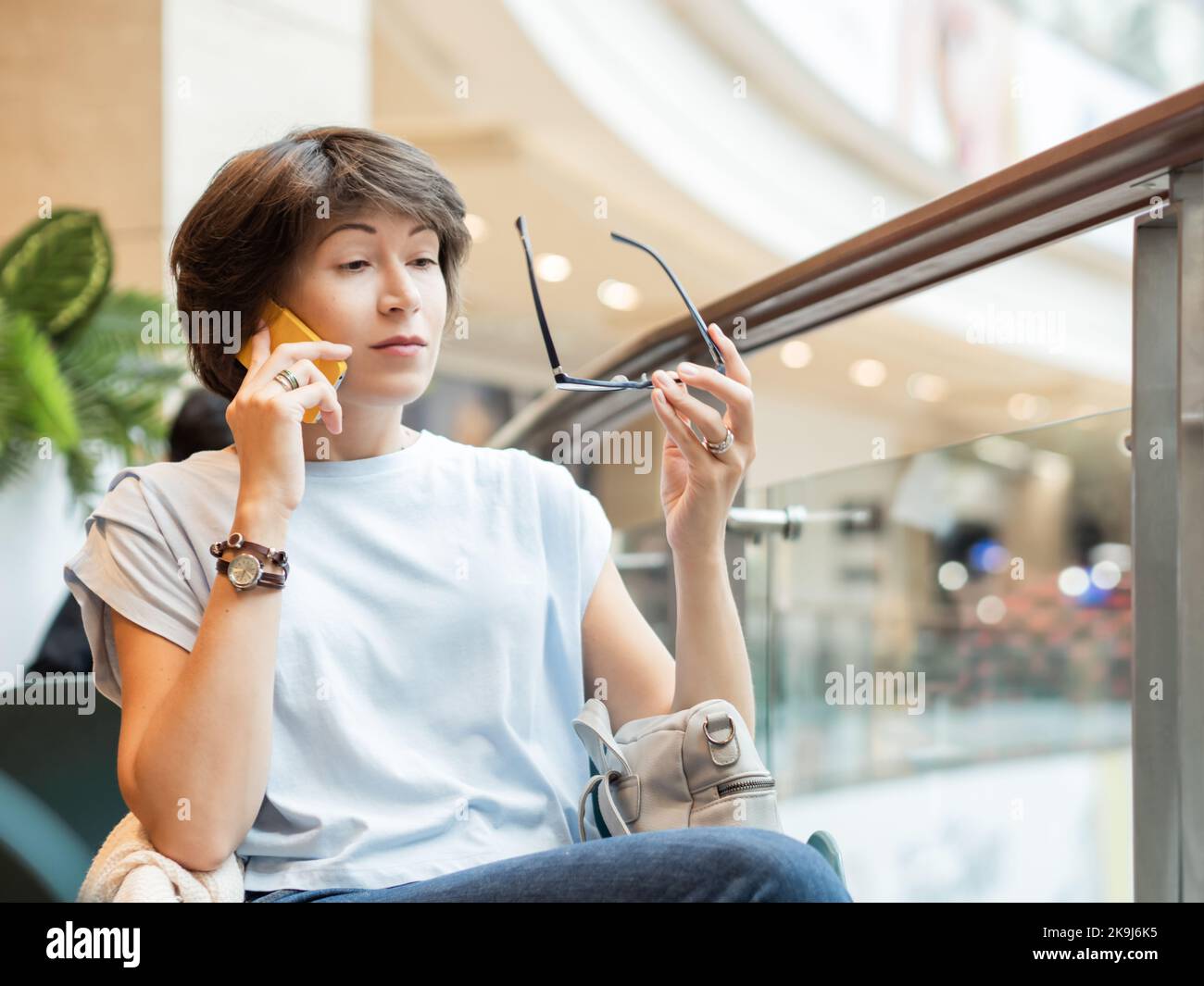 La donna sta parlando con lo smartphone e sta guardando sorprendentemente sui suoi occhiali al centro commerciale. Pausa caffè in negozio. Foto Stock