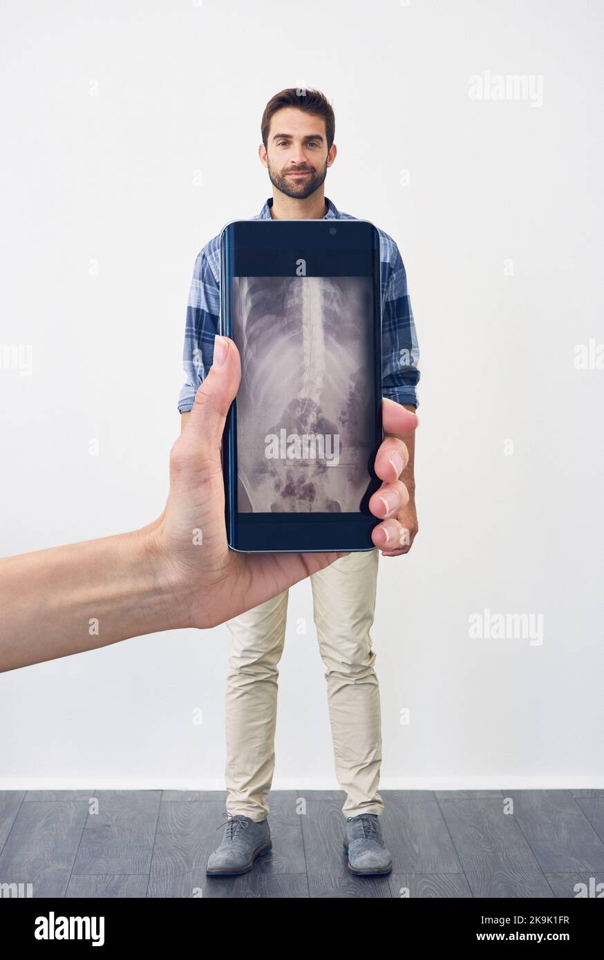 Vediamo proprio attraverso di voi: Una persona irriconoscibile che tiene un telefono che mostra una radiografia di un giovane uomo spensierato in piedi su uno sfondo grigio. Foto Stock