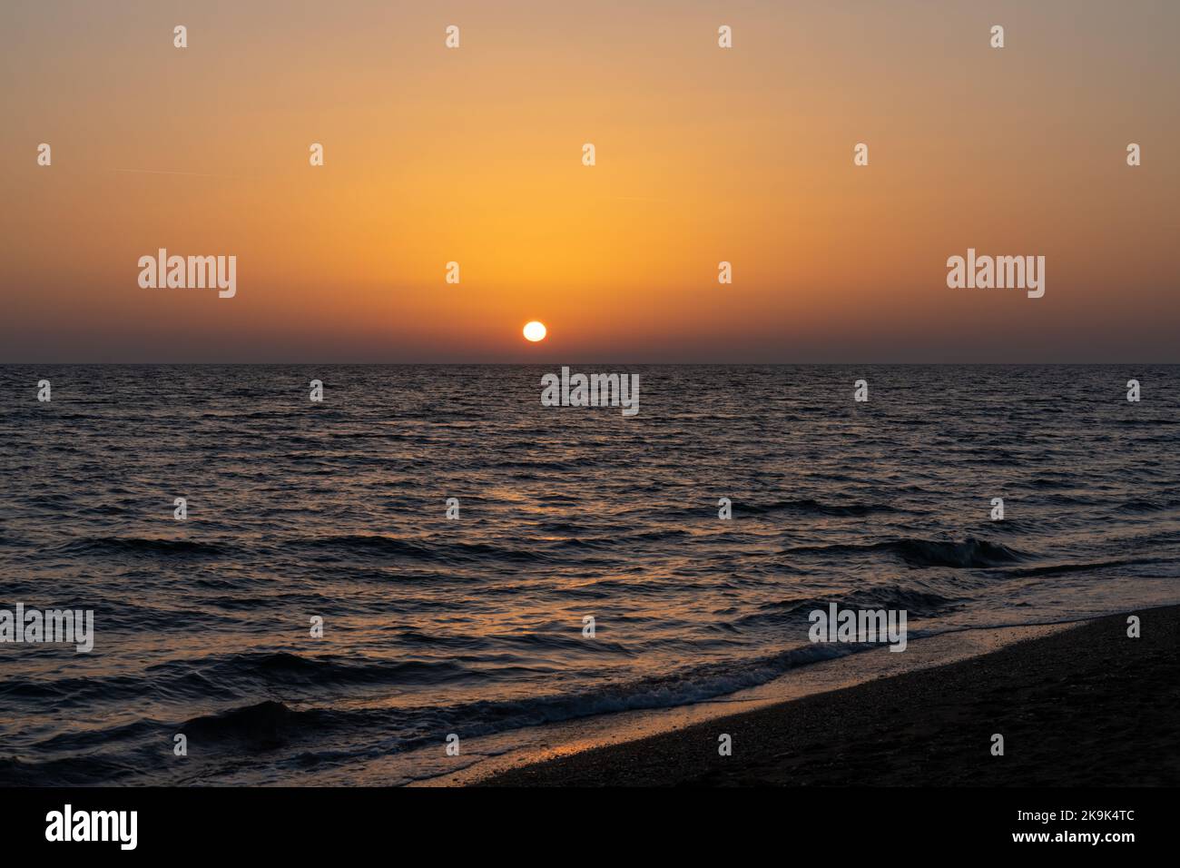 Tramonto colorato sul Mar Mediterraneo in Grecia, con calme acque oceaniche e spiaggia di sabbia scura in primo piano Foto Stock