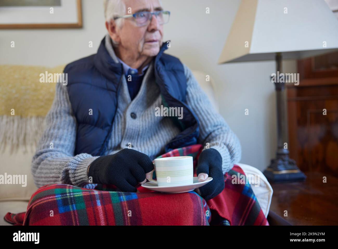 Uomo anziano che indossa vestiti extra con bevanda calda cercando di mantenere caldo a casa in crisi energetica Foto Stock