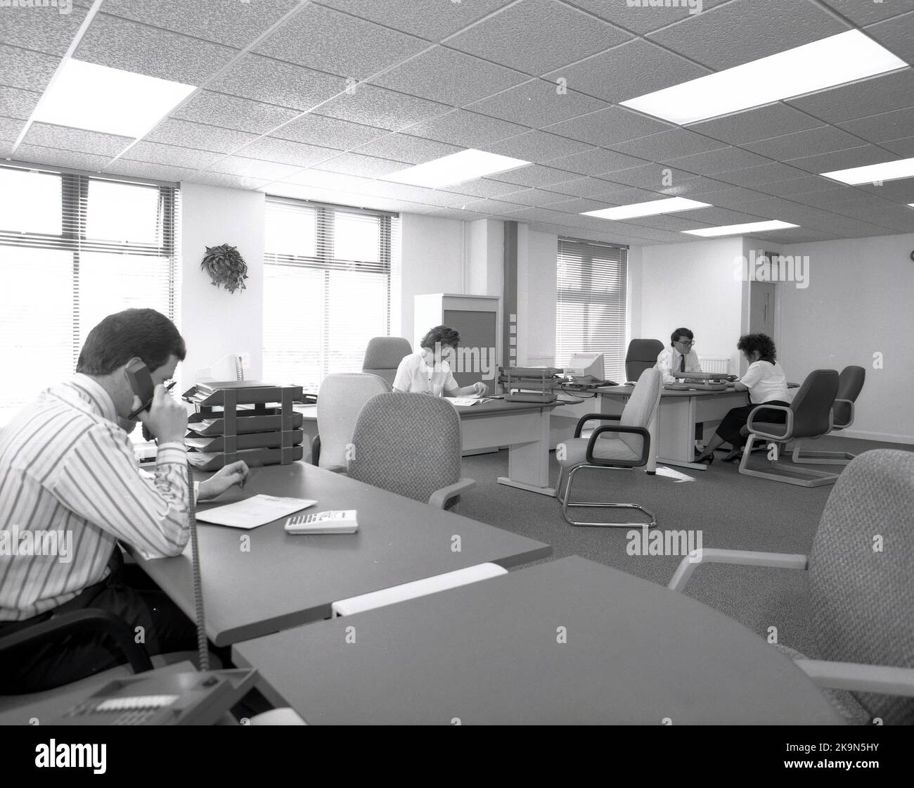 1989, storico, spazioso ufficio open space, Inghilterra, Regno Unito, i dipendenti aziendali di grandi scrivanie semicircolari che lavorano, un maschio che parla al telefono, un altro maschio che ha una discussione uno a uno con un membro del personale femminile. Un personal computer del giorno è seduto sul lato di una delle scrivanie. Foto Stock