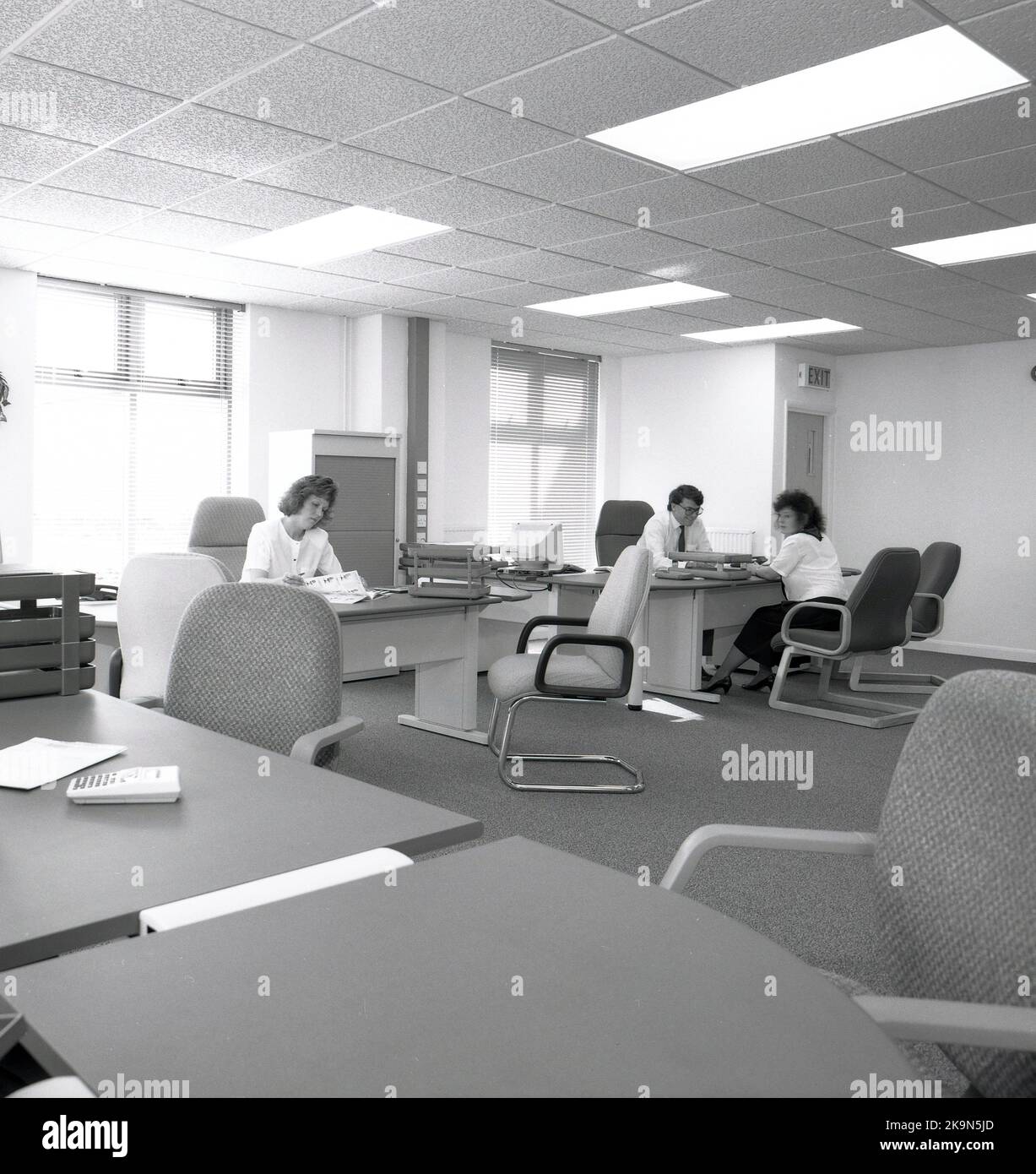 1989, storico, un maschio e una femmina che lavorano alle loro scrivanie in uno spazioso ufficio a pianta aperta, Inghilterra, Regno Unito. Grandi scrivanie semicircolari con sedie circostanti per riunioni informali - una che si svolge - e un personal computer dell'epoca in mostra. Foto Stock