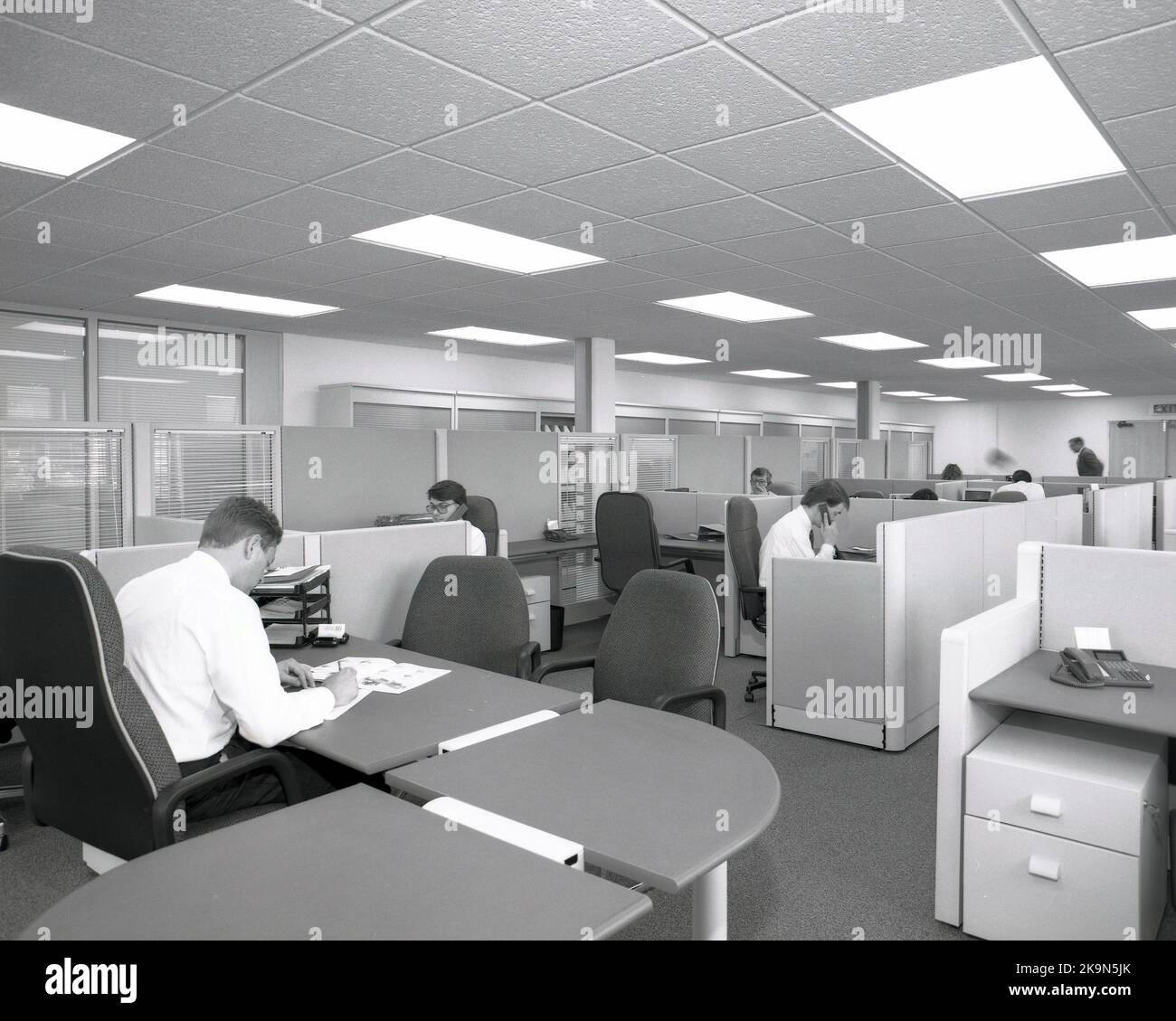 1989, dipendenti storici dell'azienda che lavorano alle scrivanie in cubicoli condivisi, in un ufficio aperto comune in quest'epoca, Inghilterra, Regno Unito, con il manager seduto in una grande scrivania semicircolare, che si affaccia sul pavimento. Foto Stock
