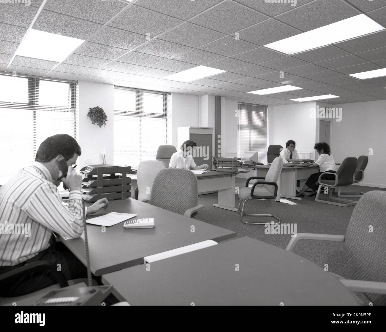 1989, dipendenti storici, di sesso maschile e femminile che lavorano alle loro scrivanie in uno spazioso ufficio a pianta aperta, Inghilterra, Regno Unito. Si possono vedere grandi scrivanie semicircolari con sedie circostanti per riunioni informali - una in corso - e personal computer dell'epoca. Foto Stock