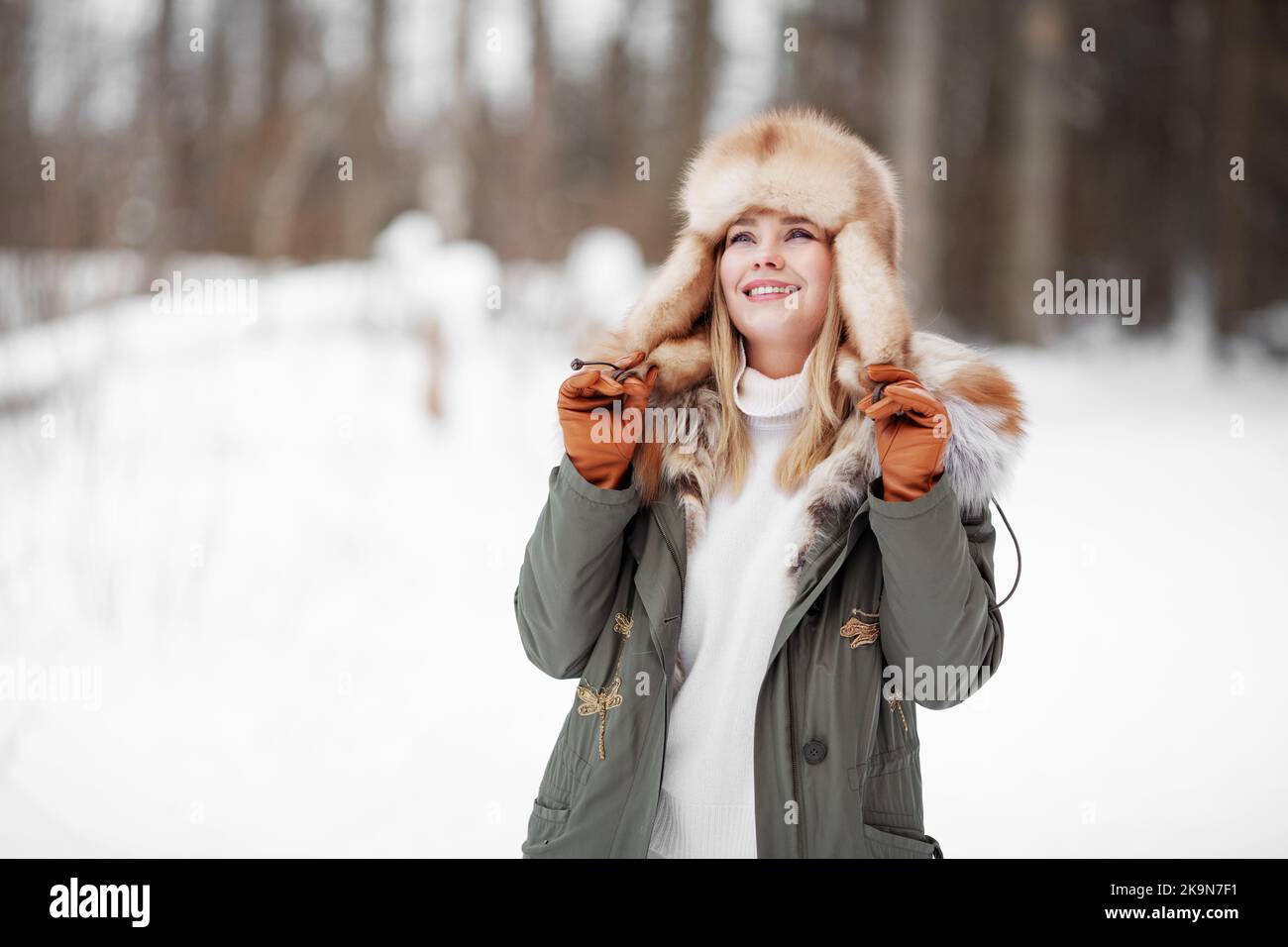 Bella donna sorridente ammirando la natura invernale nella foresta, indossando giacca di pelliccia kaki, cappello e guanti di pelle, ritratto accogliente su sfondo di neve Foto Stock