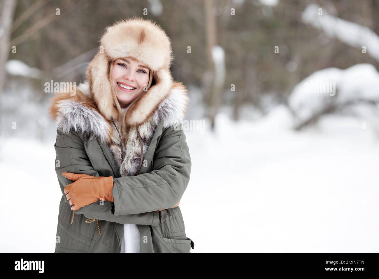 Ritratto di bella ragazza felice in piedi nella foresta di neve invernale natura. Donna sorridente che indossa abiti caldi, giacca parka invernale, cappello in pelliccia, guanti in pelle Foto Stock