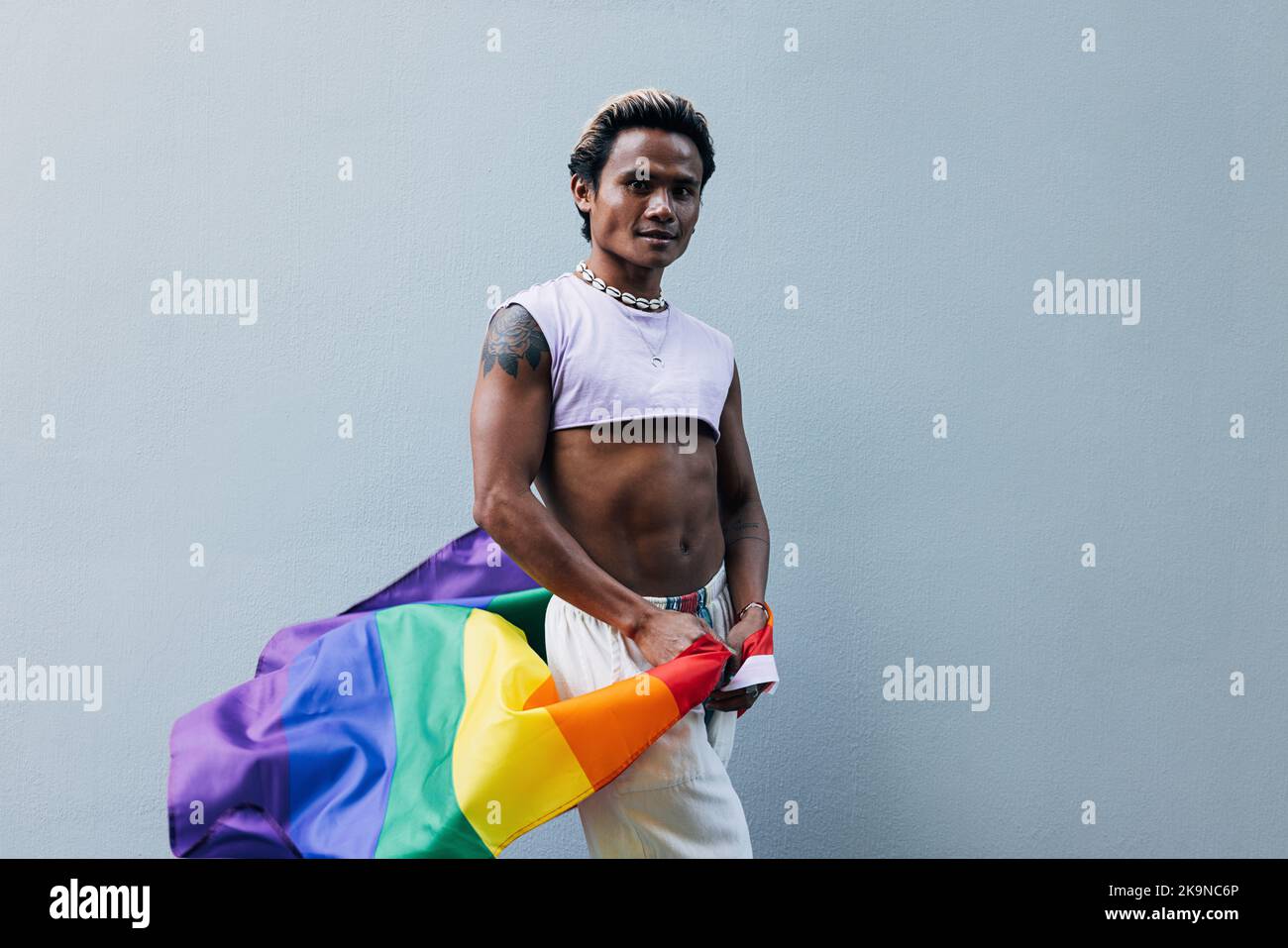 Giovane ragazzo con bandiera LGBT in posa a parete grigia all'aperto Foto Stock