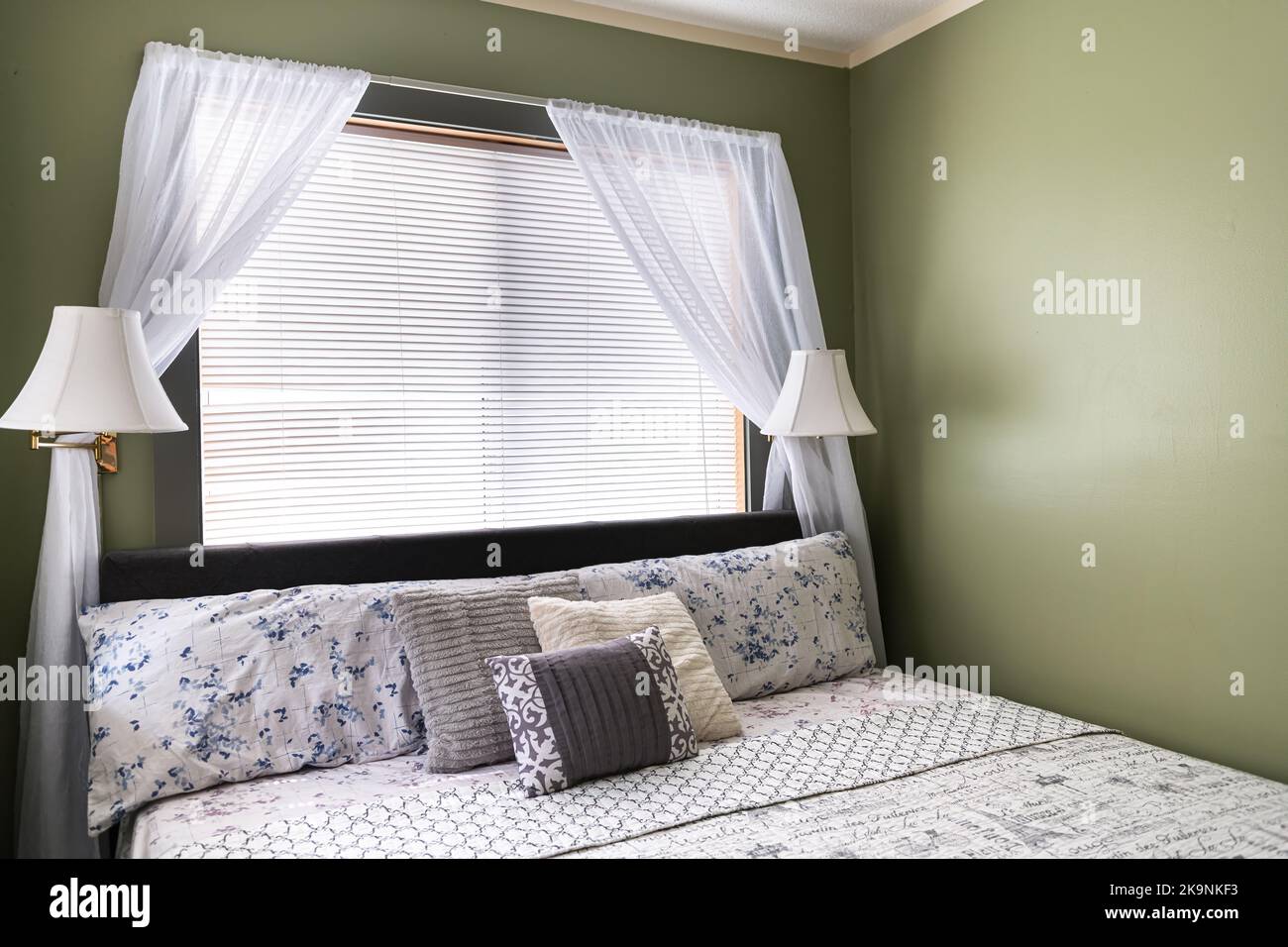 Camera da letto con lampade, cuscini con lenzuola sulla testata del letto in un moderno motel con tende bianche e pareti dipinte di verde Foto Stock