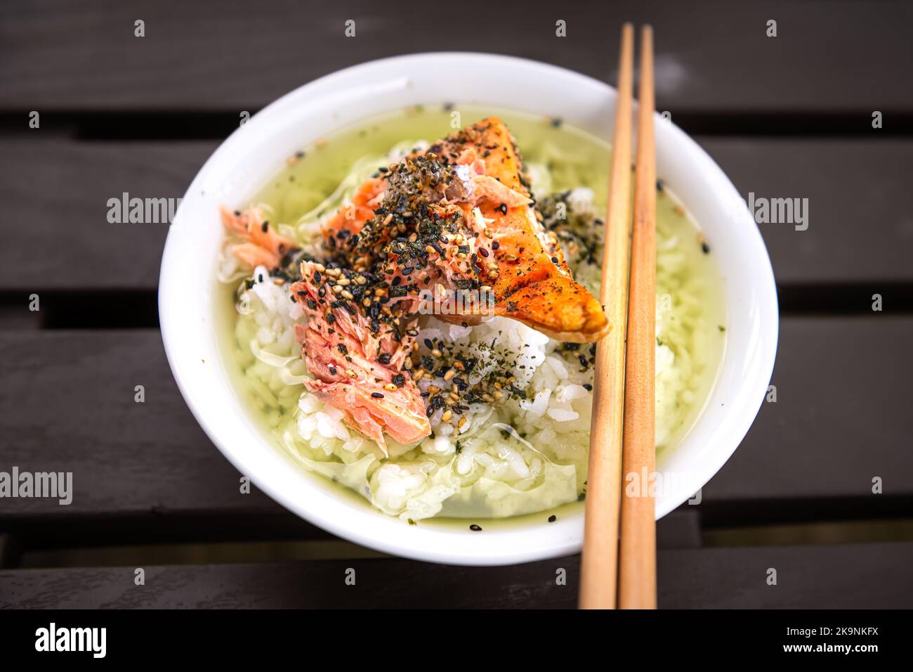 Macro primo piano del tradizionale piatto asiatico giapponese di ocazuke con salmone alla griglia, furikake di semi di sesamo di riso, nori in piatto bianco della ciotola, bastoncini Foto Stock