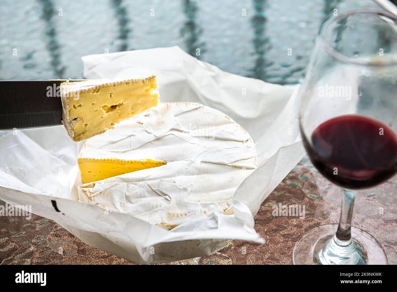 Coltello con taglio a fette di formaggio brie francese prodotto doppia crema con vino rosso in vetro su tavola da cucina macro primo piano Foto Stock