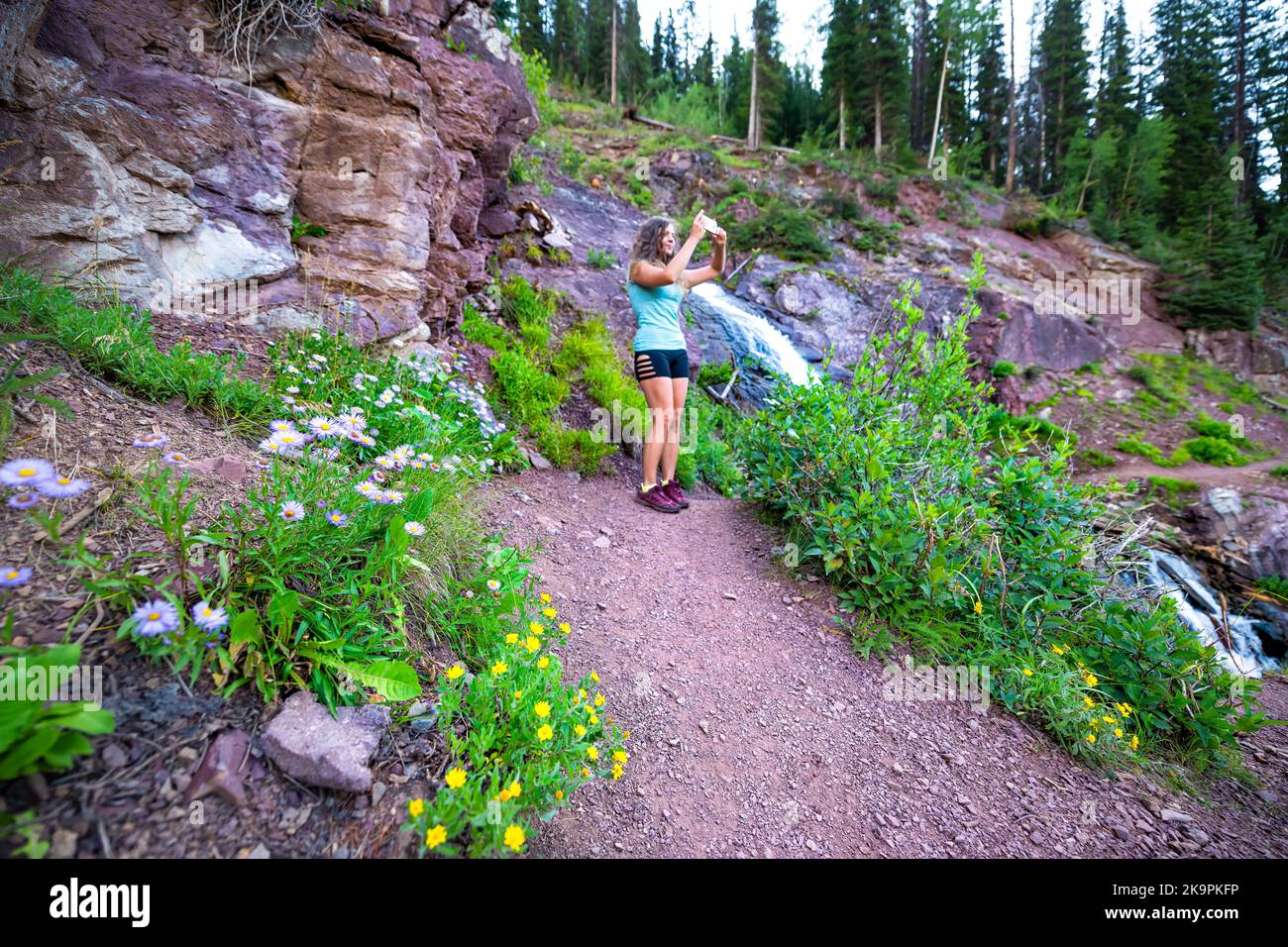 Cascata fiume sul sentiero per il lago di ghiaccio di Silverton, Colorado con percorso escursionistico fiori selvatici e giovane donna che scatta foto della natura paesaggio estivo Foto Stock