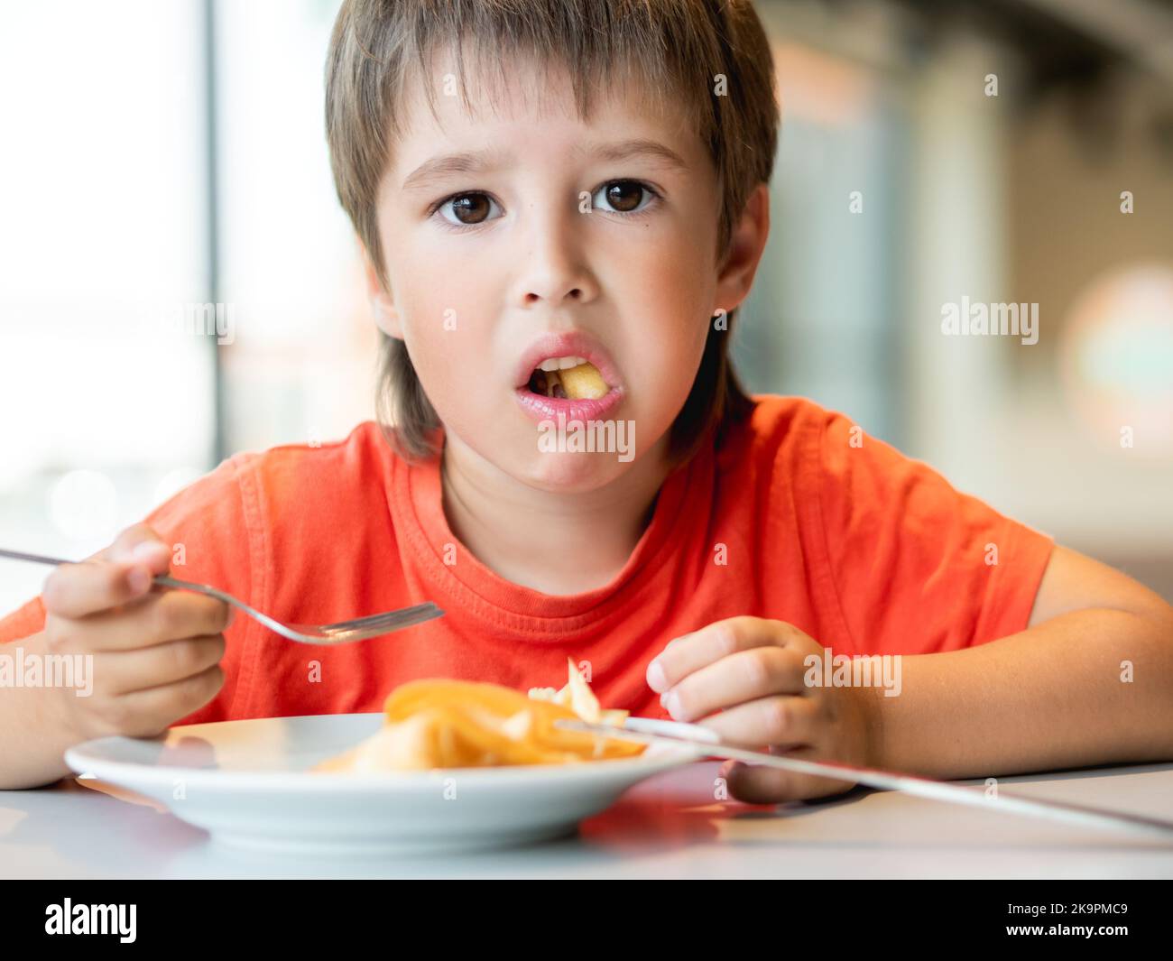 Il bambino in t-shirt rossa mangia cibo con coltello e forchetta. Ragazzo fastoso con un'espressione sconcertata sul volto. Foto Stock