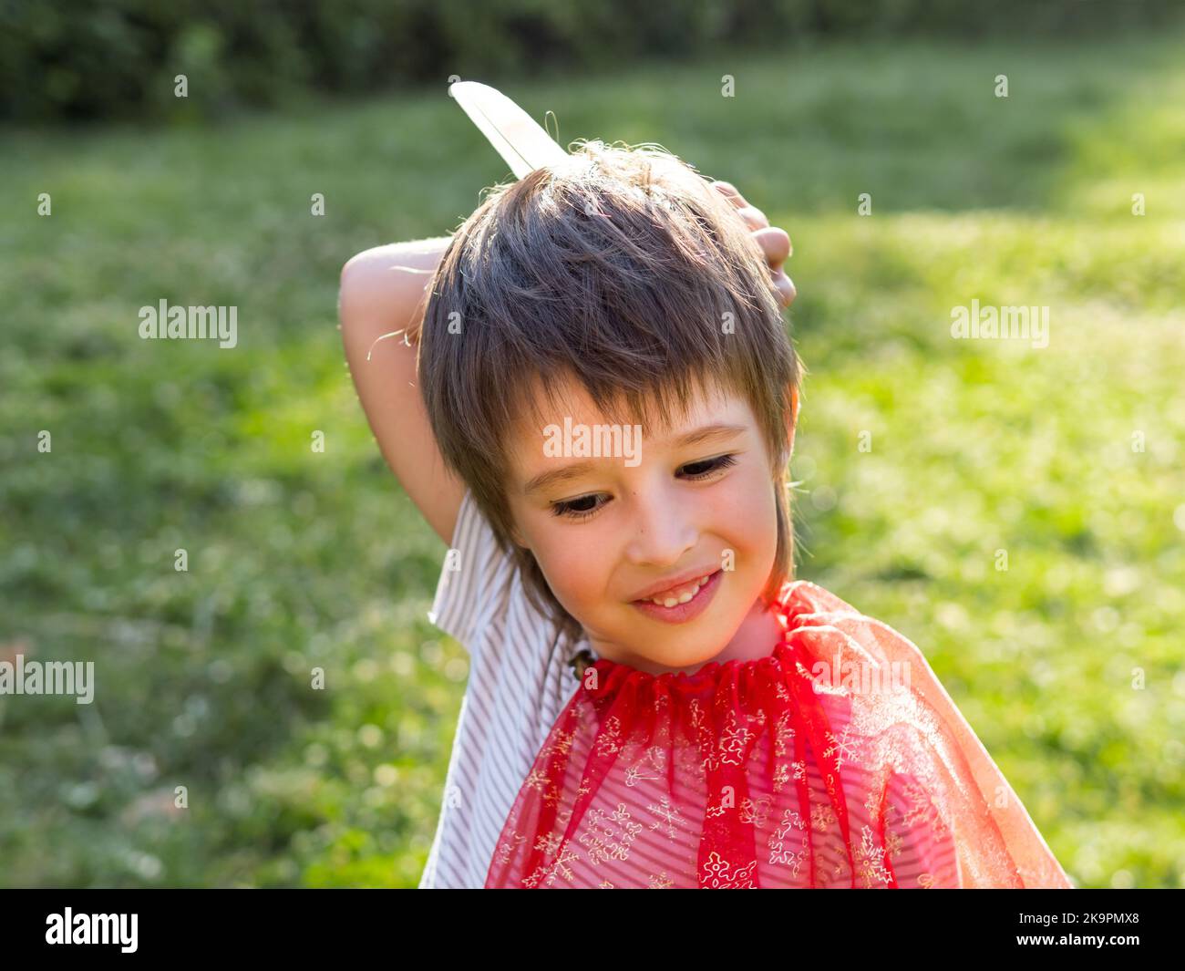 Ritratto di ragazzo sorridente che gioca americano indiano. Capretto con piume di uccello bianco e mantello rosso. Gioco di ruolo Costume. Attività ricreative all'aperto. Foto Stock