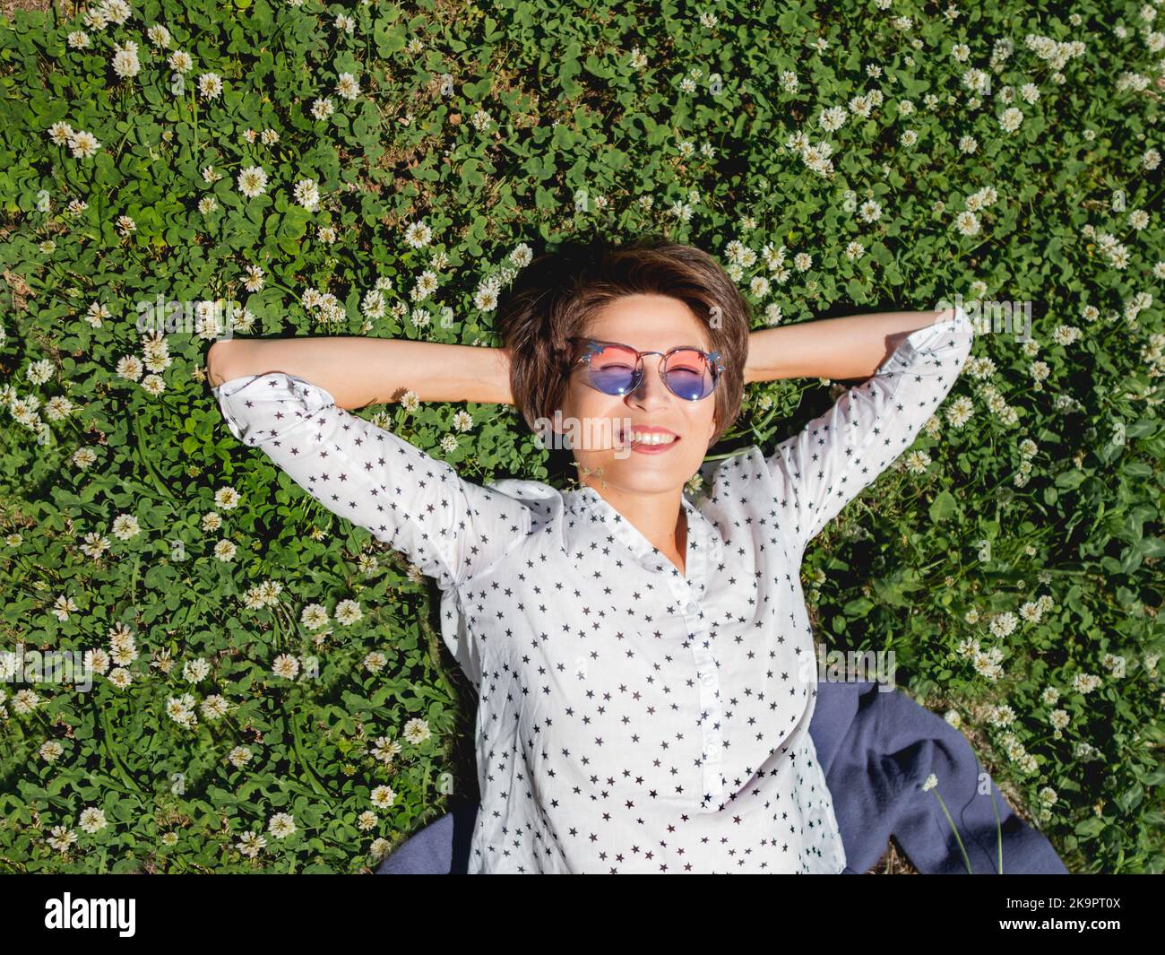Vista dall'alto sulla donna che giace tra i fiori di trifoglio sul prato nel parco urbano. Natura in città. Rilassati all'aperto dopo il lavoro. Atmosfera estiva. Foto Stock