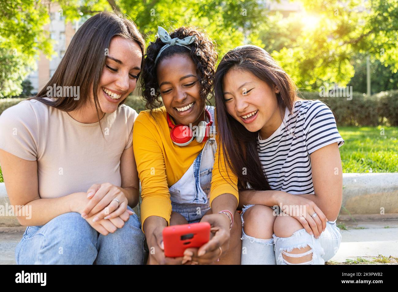 Tre giovani donne amici multirazziali che utilizzano un telefono cellulare seduto all'aperto Foto Stock