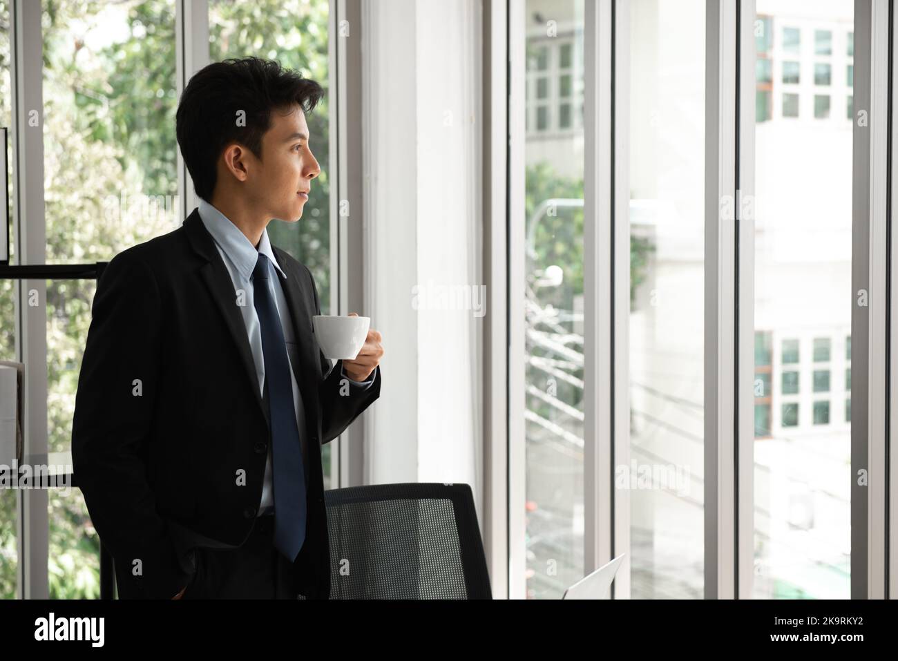 Giovane uomo d'affari premuroso che beve caffè, guardando attraverso la finestra, avendo una pausa, profondo nei pensieri, godendo di vista, in attesa dell'inizio della riunione, mak Foto Stock
