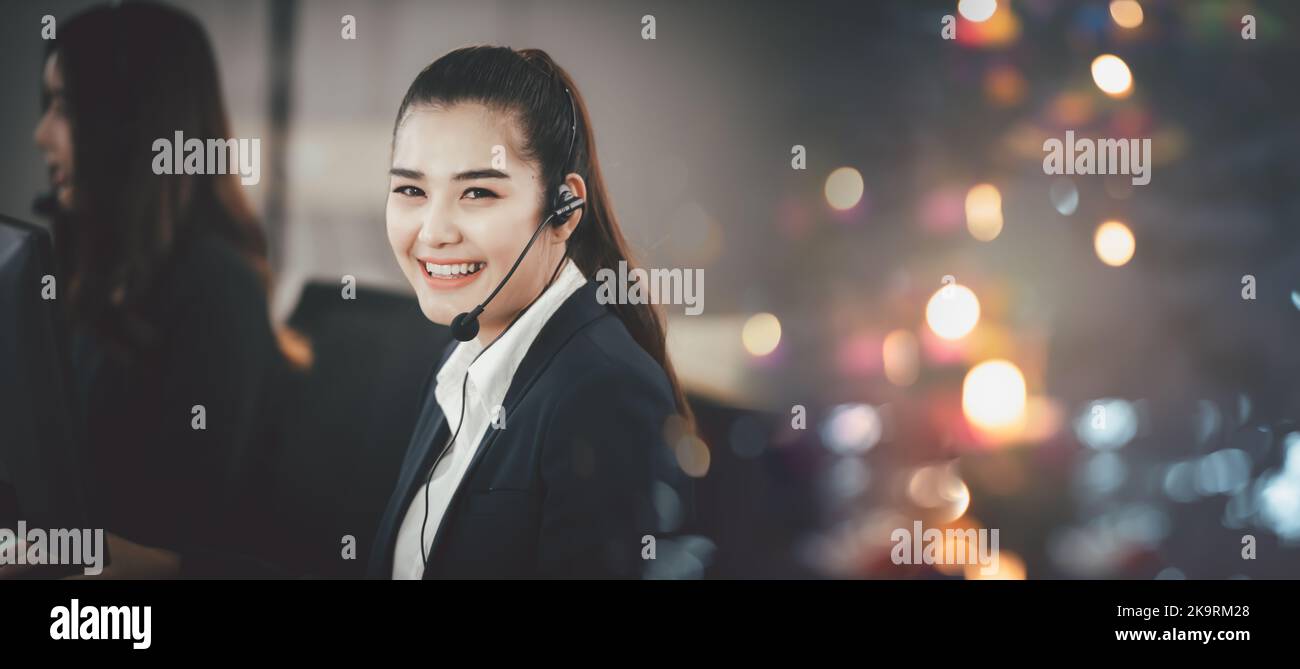 Donna asiatica in cuffia, seduta in ufficio, che lavora come operatore del call center o supporta il servizio clienti, guardando direttamente la fotocamera e sorridendo Foto Stock