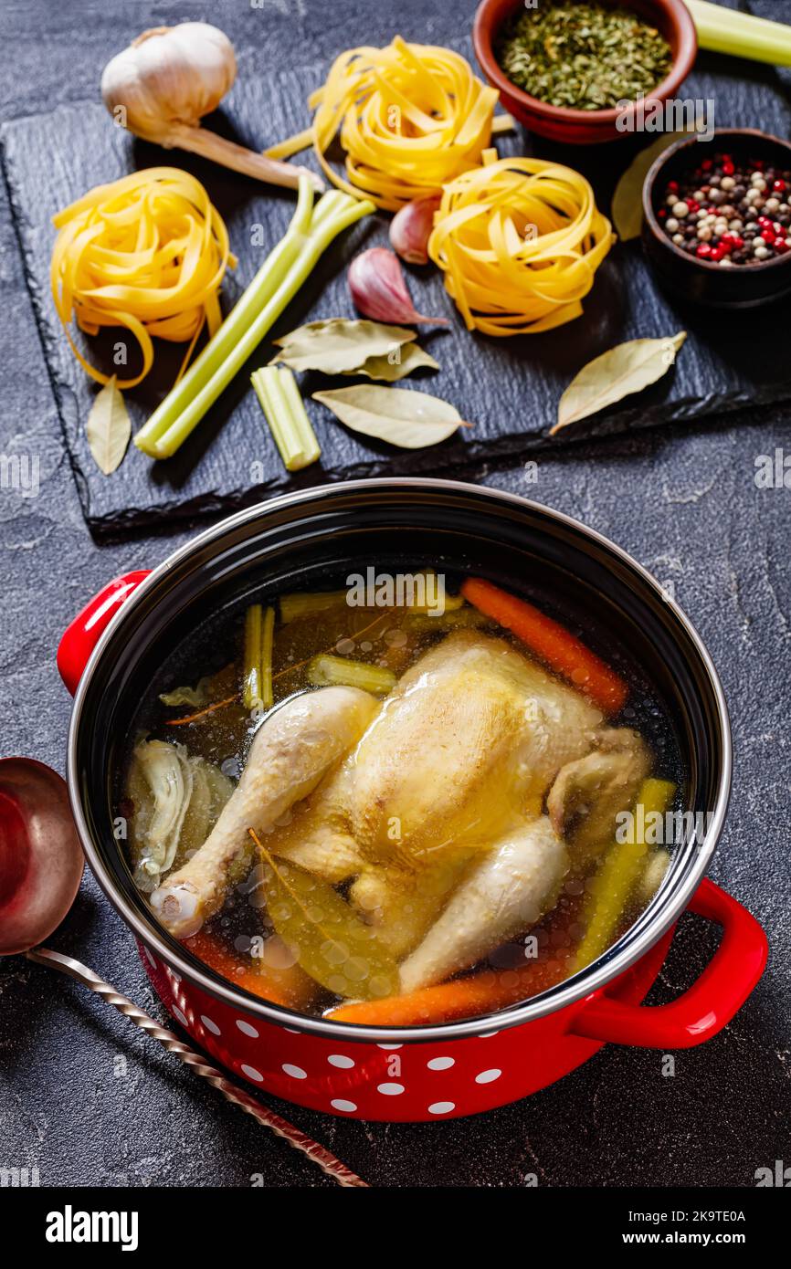 brodo di pollo cotto con pollo intero, sedano, cipolla e carota in pentola rossa con ingredienti e tagliatelle su tavolo di cemento, vista verticale Foto Stock