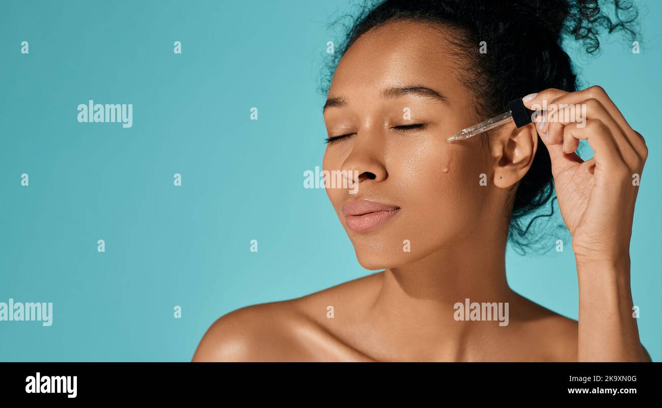 cura della pelle del viso con olio e siero essenziali. Donna africana felice che applica olio essenziale sul viso per una pelle idratata con contagocce per pipette, isolata Foto Stock