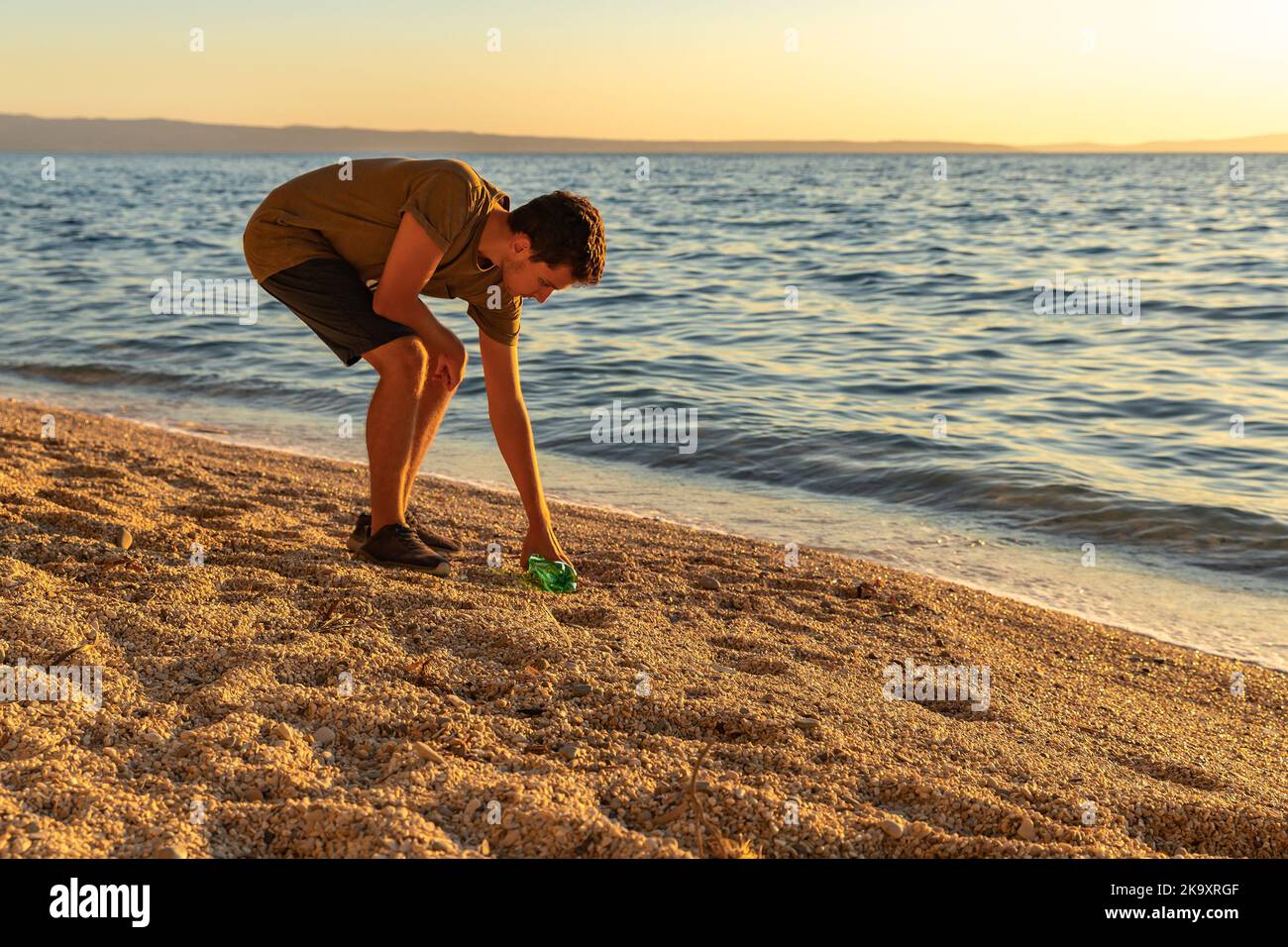 Giorno della terra. Pulire la spazzatura sulla costa adriatica. Un ragazzo che raccoglie una bottiglia di plastica sulla spiaggia. Tramonto. Il concetto di conservazione dell'ecologia Foto Stock