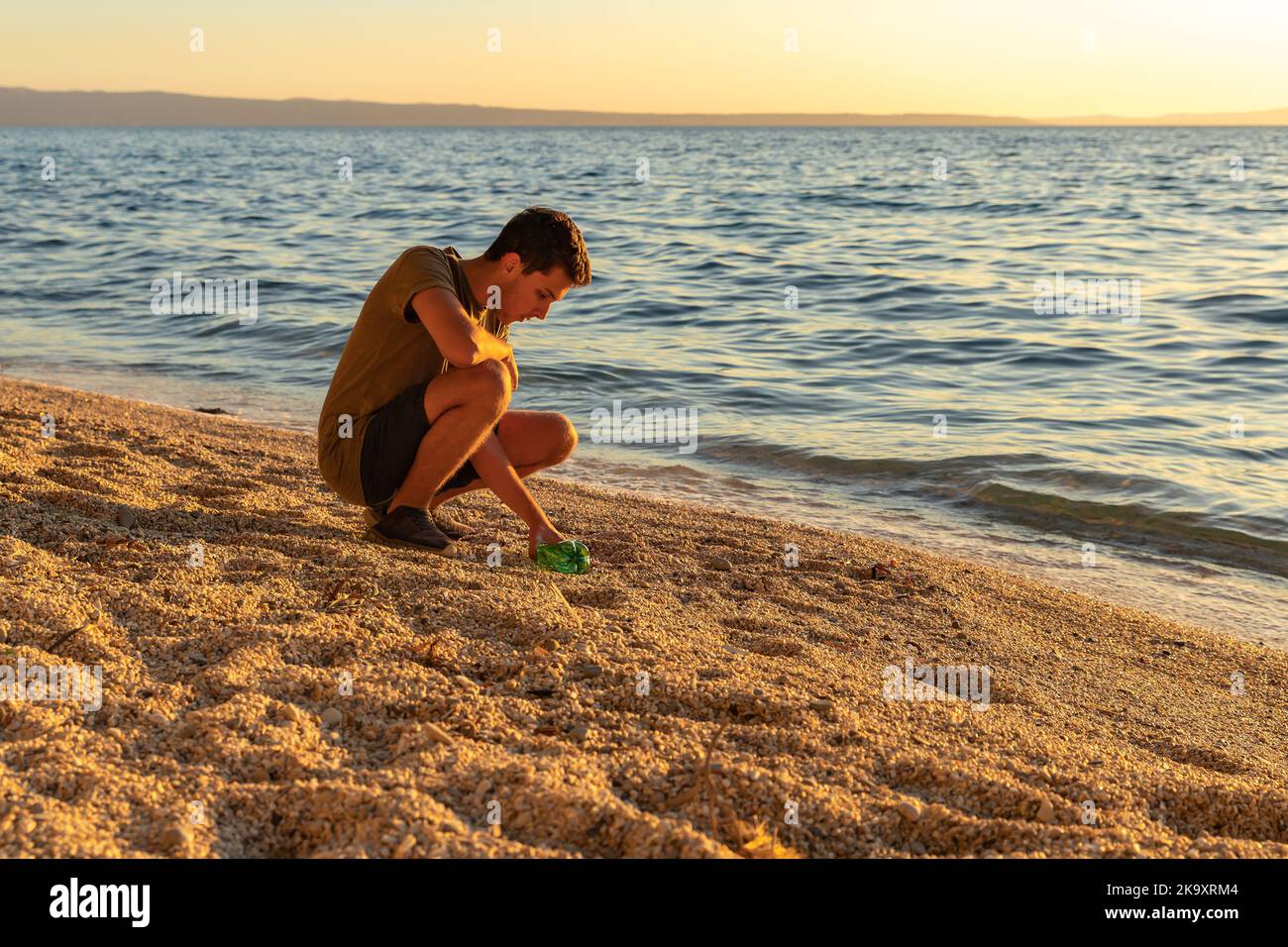 Giorno della terra. Pulire la spazzatura sulla costa adriatica. Un ragazzo che raccoglie una bottiglia di plastica sulla spiaggia. Tramonto. Il concetto di conservazione dell'ecologia Foto Stock