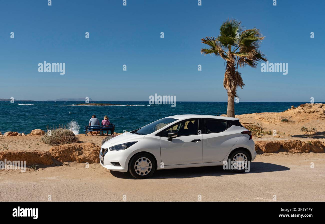 Malia, Creta, Grecia. 2022. Coppia seduta su una panchina sul lungomare di Malia, Creta con una piccola auto a noleggio in una giornata estiva. Foto Stock