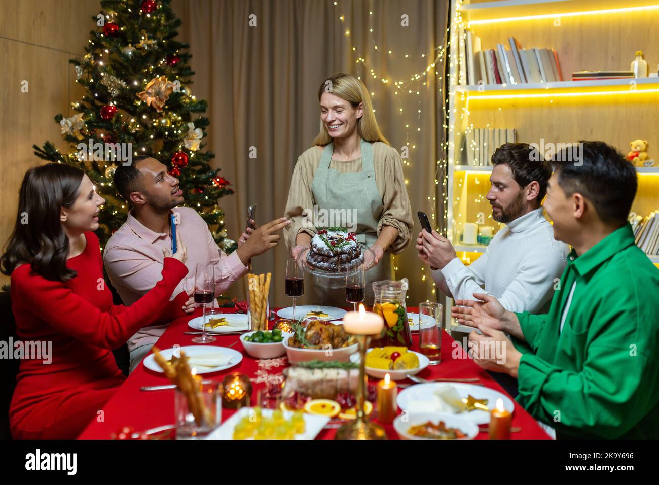 Diversi amici in visita per Natale festeggiamo le vacanze di Capodanno cenando e gioendo divertirsi, gruppo di persone che mangiano e bevono vino al tavolo a casa vicino all'albero di Natale. Foto Stock