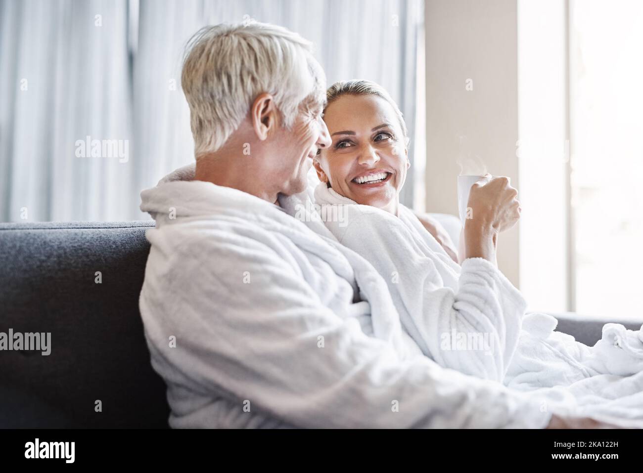 Ci meritiamo di essere viziati, ora e poi. Una coppia allegra di mezza età che si rilassa insieme indossando gli accappatoi e sedendosi su un divano all'interno di un centro benessere Foto Stock