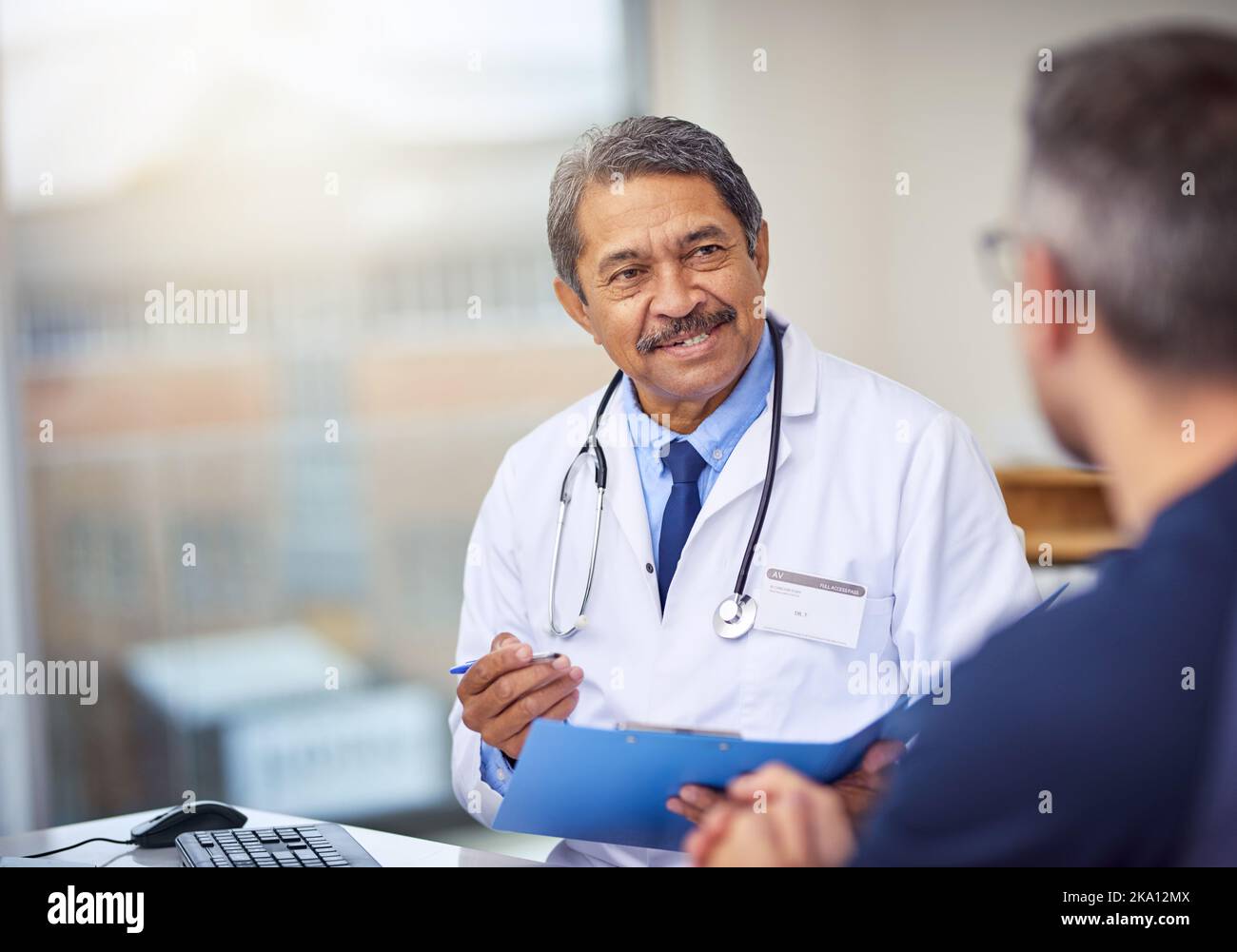 Quando ha fatto questo inizio di dolore. Un medico maschio maturo allegro seduto alla sua scrivania mentre consultandosi un paziente all'interno di un ospedale durante il giorno. Foto Stock