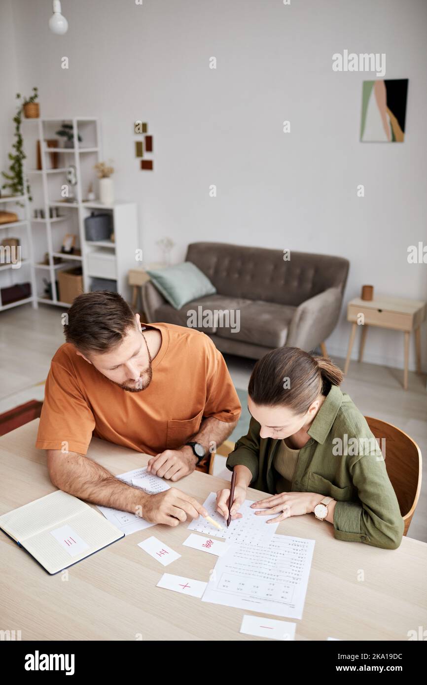 Vista dall'alto di un giovane tutor che spiega un nuovo argomento alla lezione a casa degli studenti mentre indica un documento con compiti o dati Foto Stock