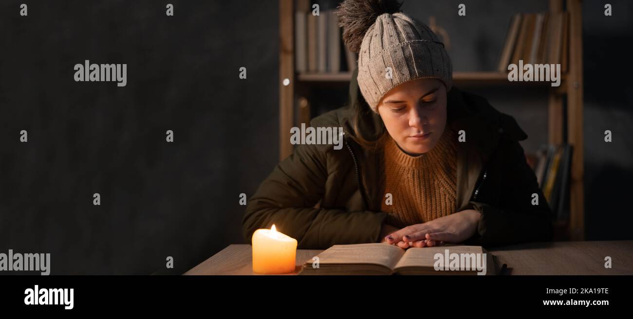 Giovane donna con abiti caldi sentendo il freddo dentro casa, casa scura senza elettricità, studente che studia utilizzando candele. Concetto di non riscaldamento e. Foto Stock