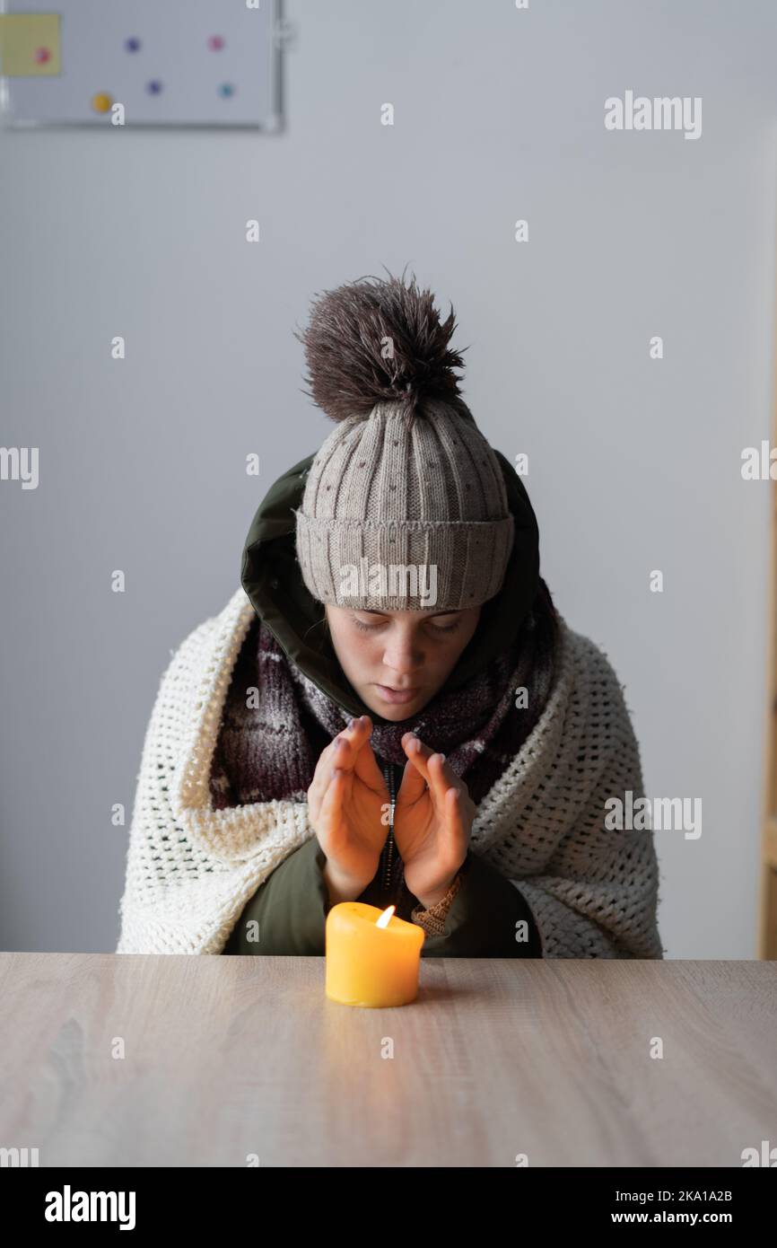 Donna malata stanca che si gelava a casa, seduta al tavolo che scalda le mani di una candela che brucia. Donna con problemi di riscaldamento a casa sensazione di freddo. Non c'è riscaldamento Foto Stock
