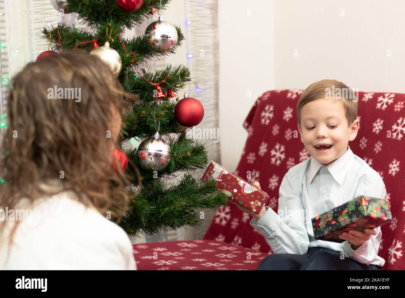 Mamma dà i suoi regali del figlio di sette anni in scatole per il nuovo anno contro lo sfondo di un albero di Natale a casa. Messa a fuoco selettiva. Verticale Foto Stock