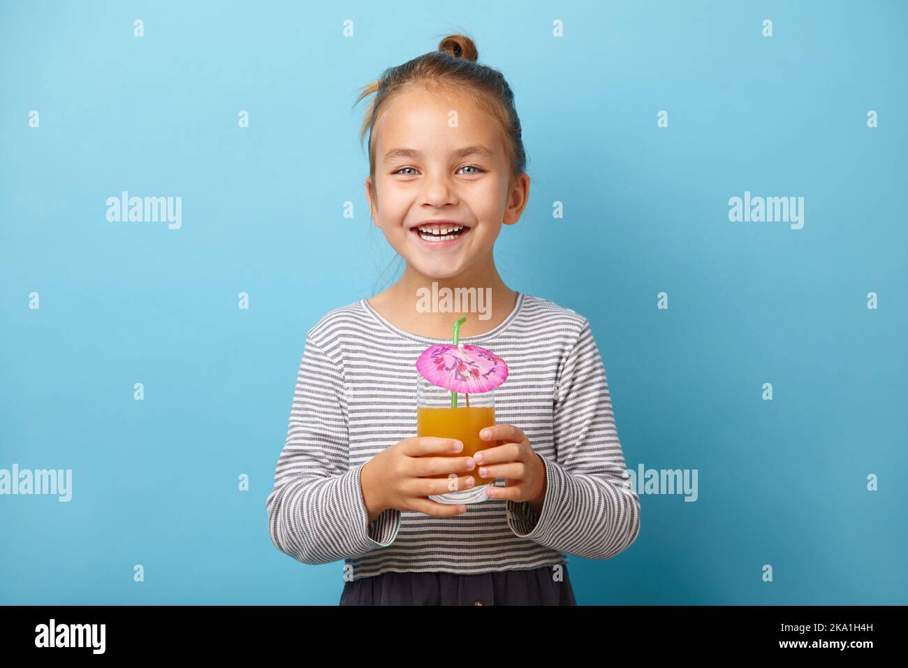 Allegra bambina beve succo d'arancia e ride, si trova su sfondo isolato blu. Foto Stock