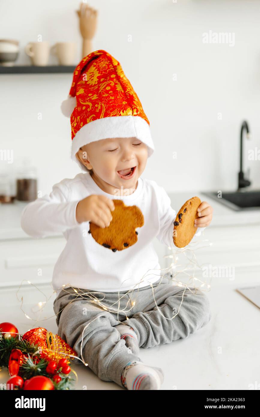 Un ragazzino si sveglia al mattino e mangia i biscotti di Natale con il latte in cucina ridendo e felice. Mattina di Natale. Foto Stock