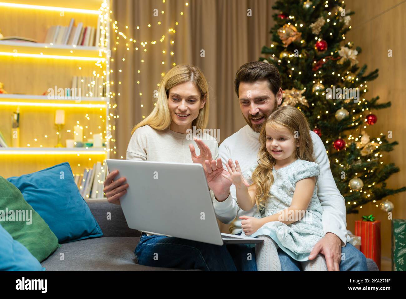 Quarantena per le vacanze. Una famiglia giovane, un uomo, una donna e una bambina sono seduti insieme sul divano di casa vicino all'albero di Natale. Parlano e salutano i loro genitori tramite videochiamata da un laptop. Foto Stock