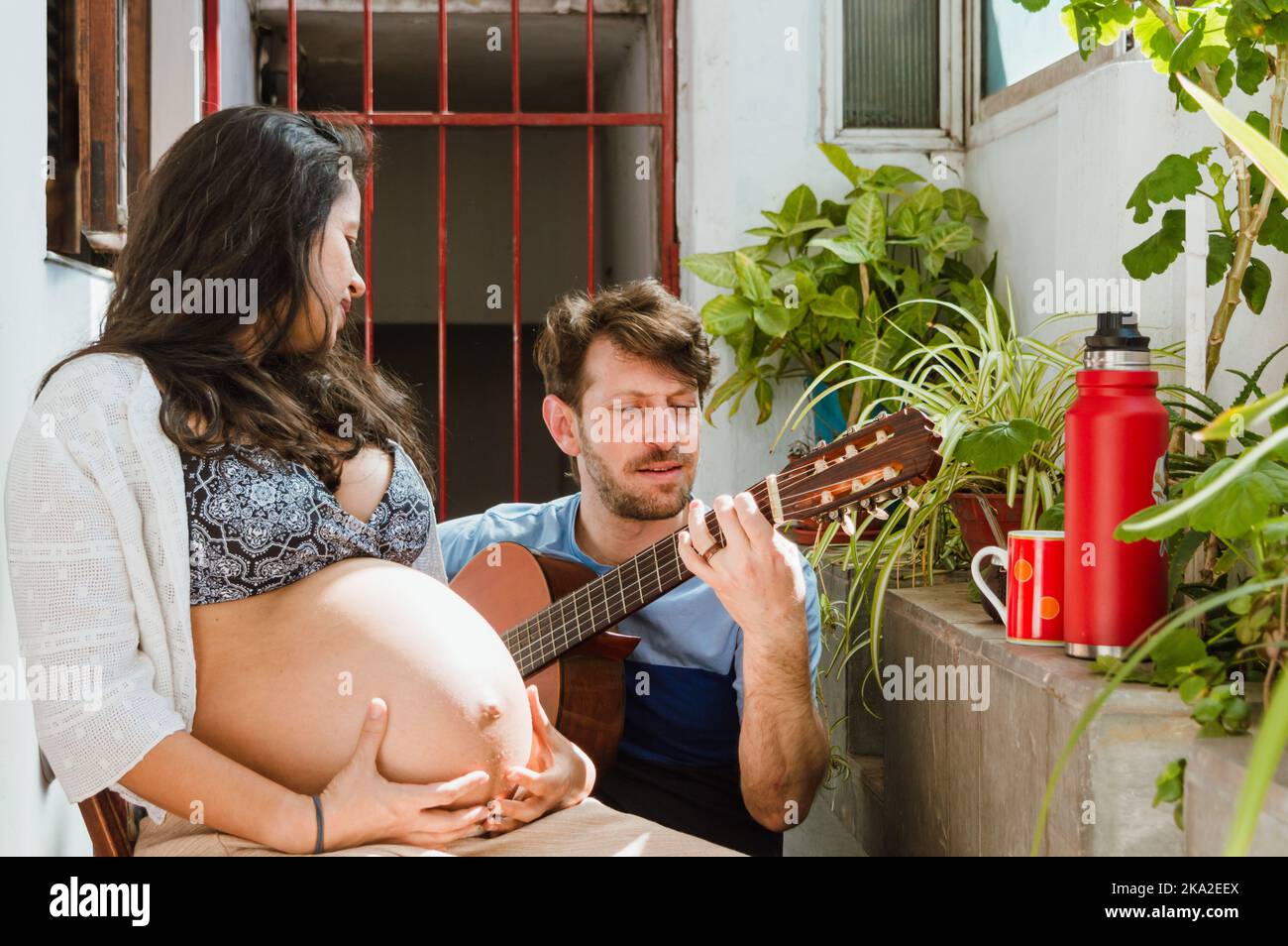 il giovane argentino con capelli corti e barba si accoccolò a casa e mostra come suona la chitarra a sua moglie una donna di etnia brasiliana Foto Stock