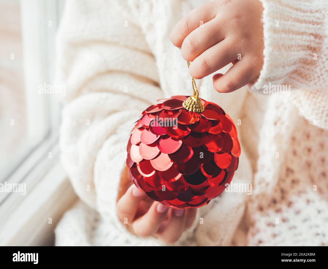 Capretto con sfera decorativa rossa per albero di Natale. Ragazzo in maglia a cavetto oversize. Vestito accogliente per un clima sereno. Lo spirito delle vacanze invernali. Anno nuovo Foto Stock
