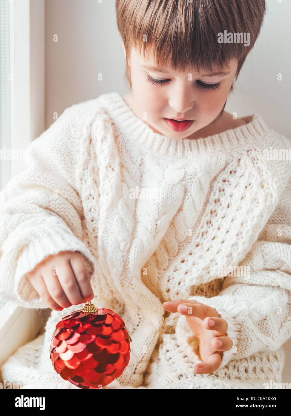 Capretto con sfera decorativa rossa per albero di Natale. Ragazzo in maglia a cavetto oversize. Vestito accogliente per un clima sereno. Lo spirito delle vacanze invernali. Anno nuovo Foto Stock