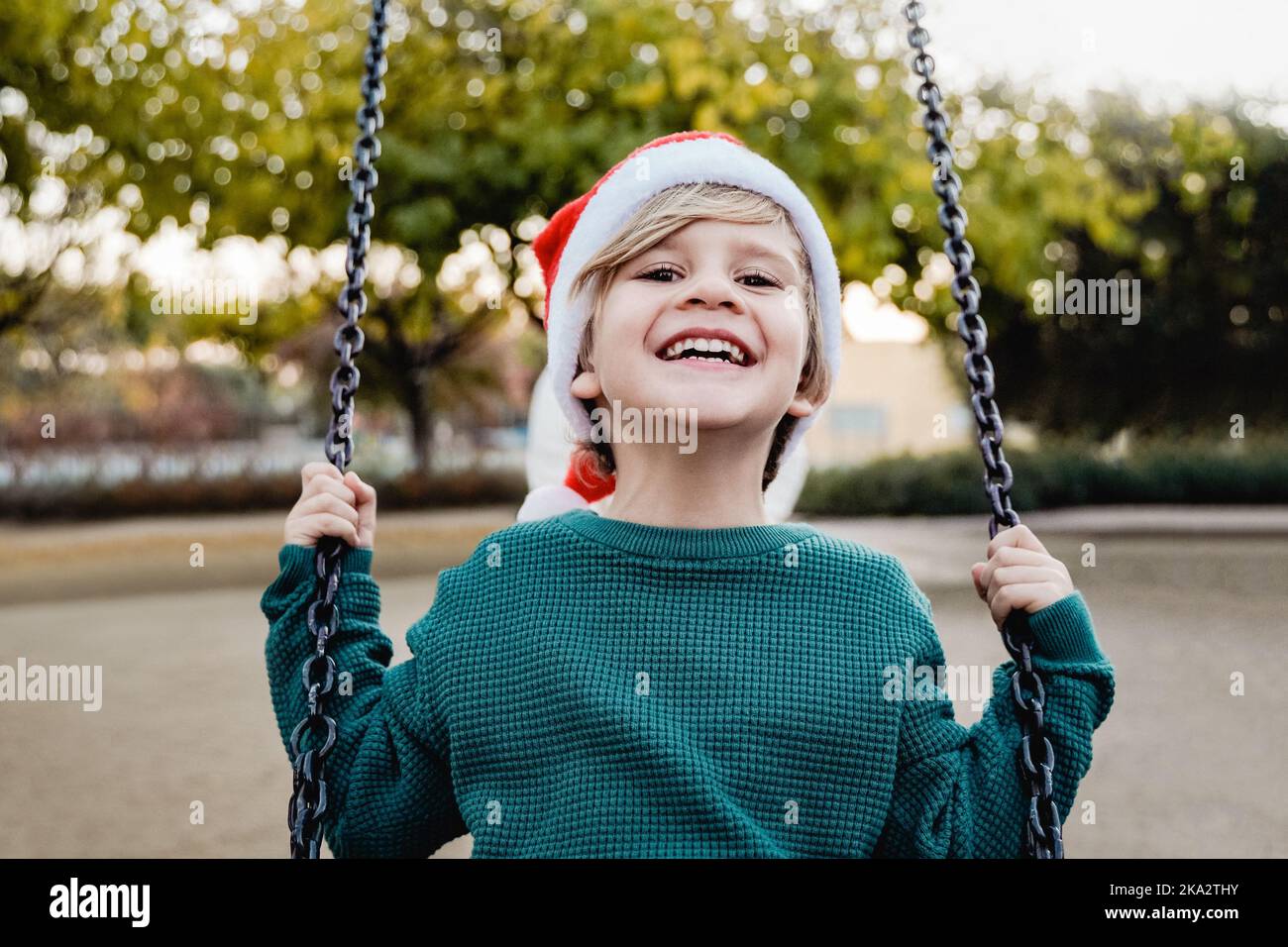 Bambino felice che indossa il cappello di Babbo Natale che si diverte nell'altalena all'aperto al parco della città - concetto di Natale - fuoco sul fronte Foto Stock