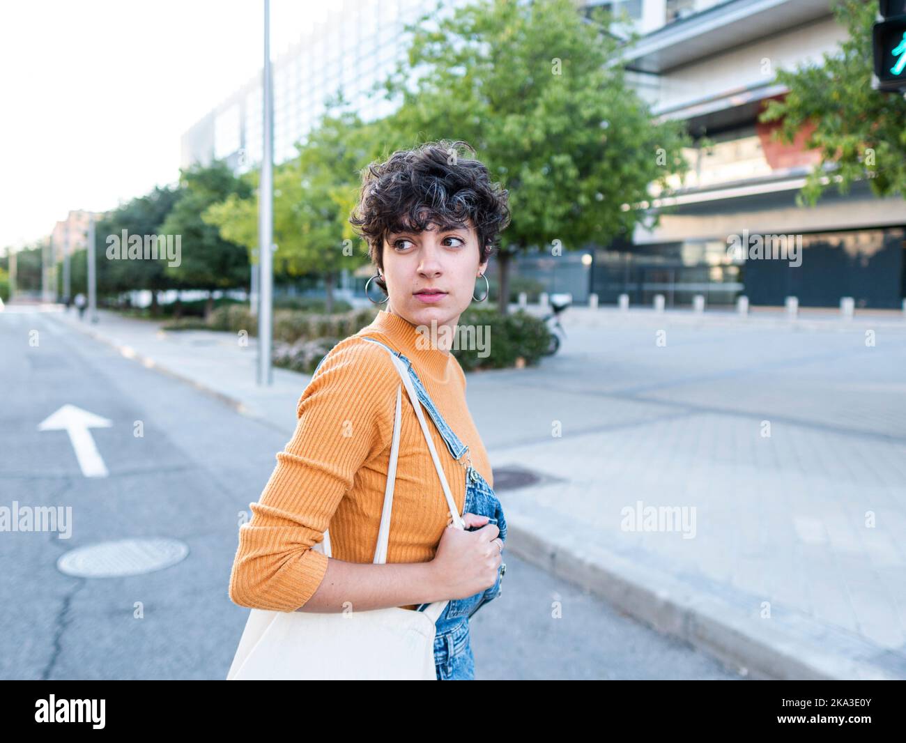 Vista laterale di una giovane donna con capelli scuri corti in abiti casual camminando sulla strada vicino a un edificio urbano a più piani e guardando lontano Foto Stock