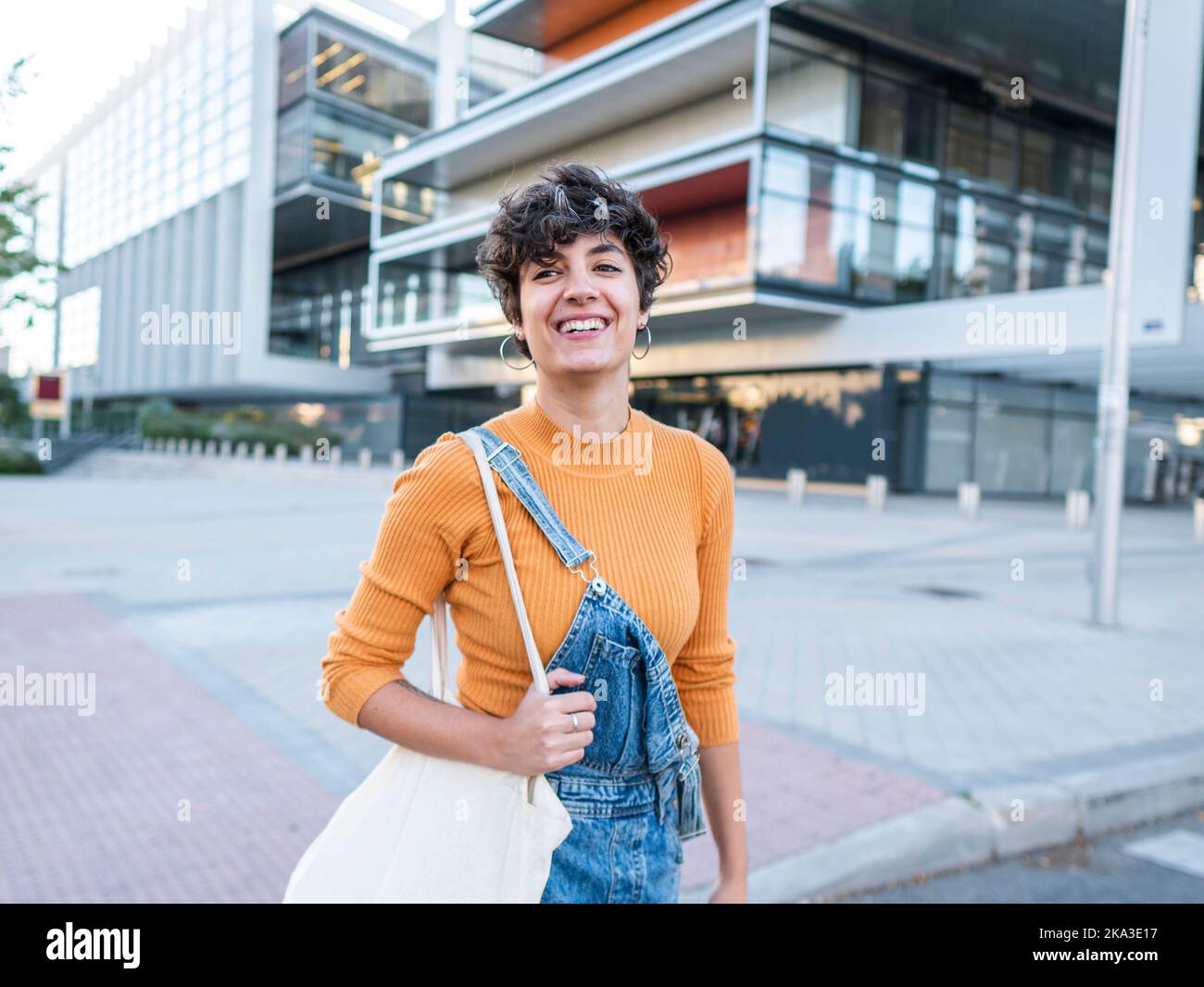 Felice giovane donna con capelli corti scuri in abiti casual in piedi sulla strada vicino a edificio urbano a più piani e guardando lontano Foto Stock