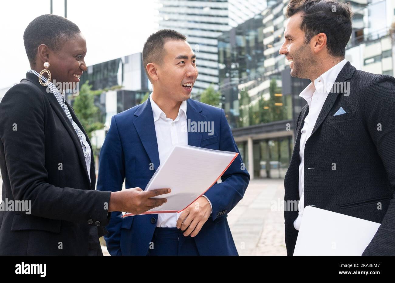 Colleghi multietnici in abiti formali in piedi per strada e tenere cartelle mentre si discute di progetti aziendali e si guarda l'un l'altro vicino a più persone Foto Stock