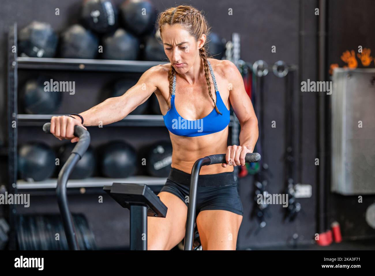 Atleta muscoloso focalizzato facendo esercizio su air bike contro scaffali con pesi durante l'allenamento funzionale in palestra Foto Stock